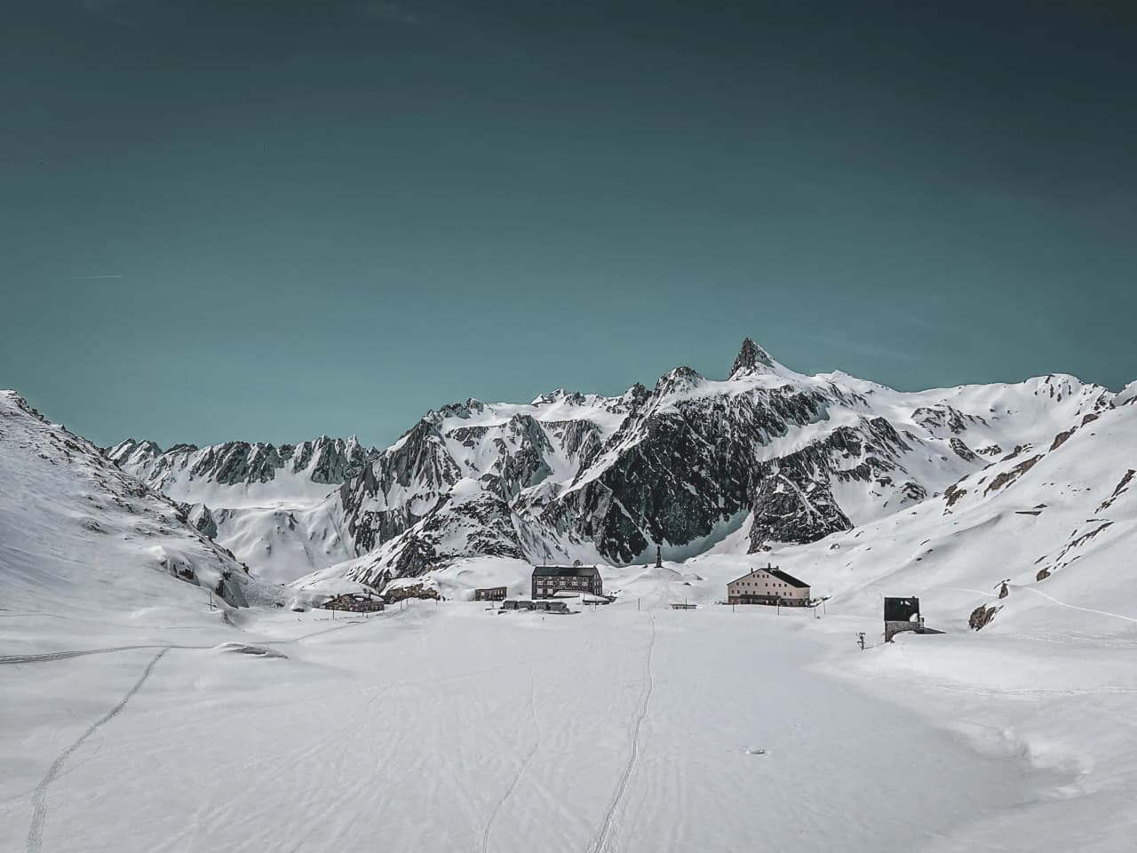 A snow-covered alpine landscape with mountain huts, bordered by majestic peaks under a blue sky.