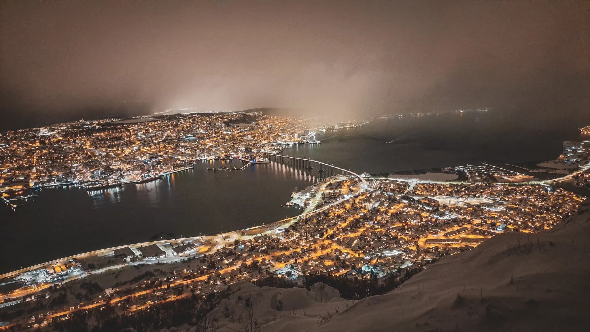 Night view of the illuminated town on the edge of a fjord in Norway, under a starry sky.