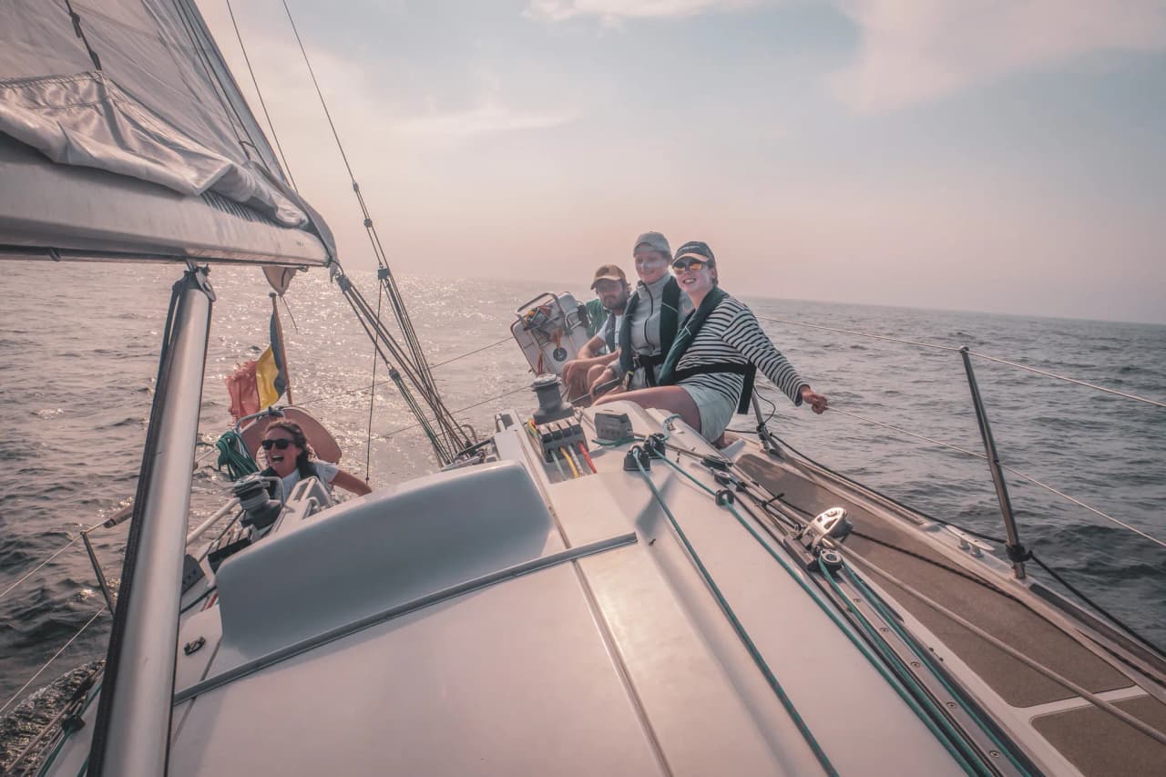 Vier lachende mensen op een zeilboot, met de zon die schittert op de oceaan.
