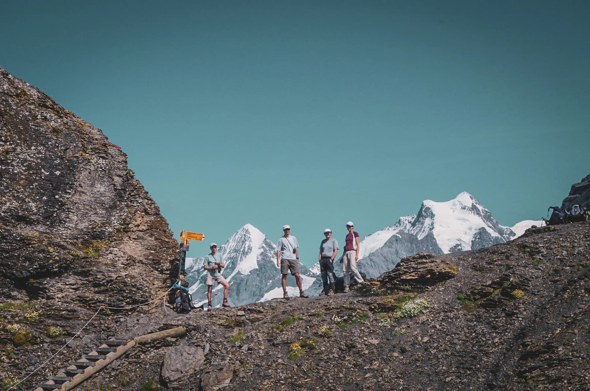 A group of five hikers are standing on rocky ground, surrounded by majestic snow-capped mountains in the background. One of the hikers appears to be taking photographs.