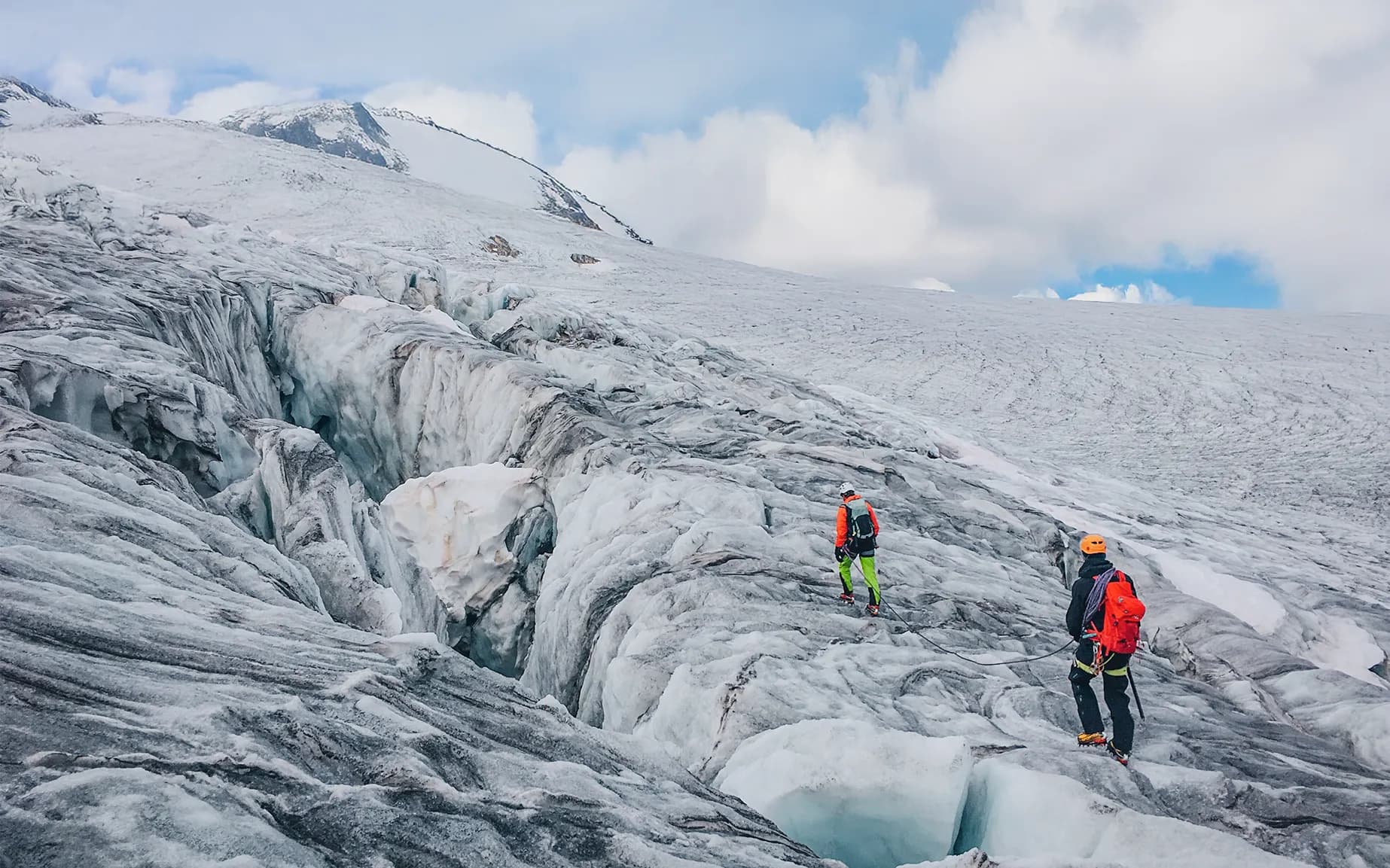 A monumental glacier with deep crevasses and complex ice formations occupies the foreground. Two climbers, one in an orange jacket and the other in black with a red rucksack, are walking along the glacier.