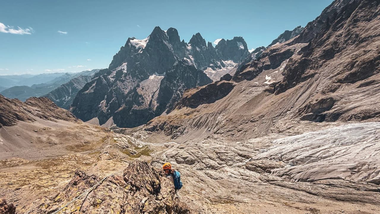 Un alpiniste grimpe une montagne majestueuse dans les Écrins, sous un ciel dégagé et ensoleillé.