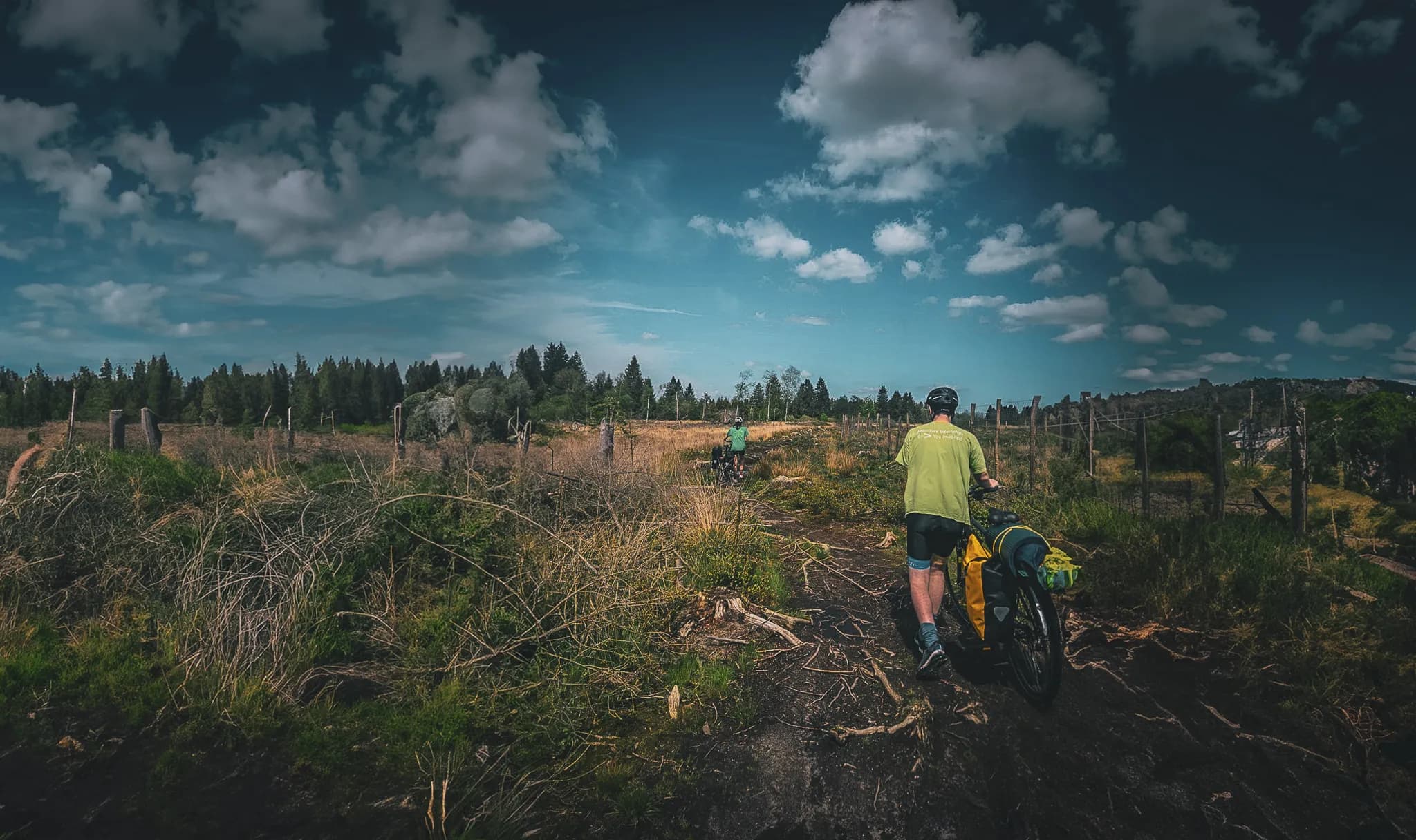 A dirt track crossing a rural landscape, bordered by fields and vegetation. Two cyclists, one carrying a yellow rucksack, are walking along the track, while a third cyclist can be seen further back.
