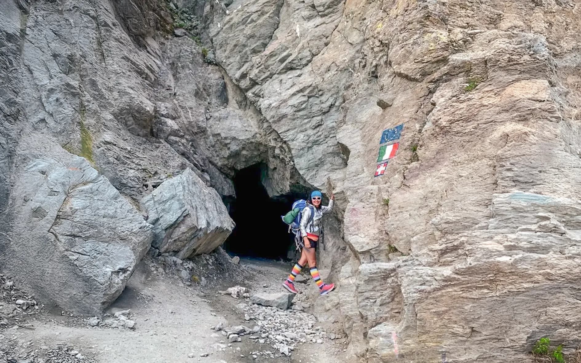 Happy hiker near a mountain cave, displaying flags and a striking landscape.