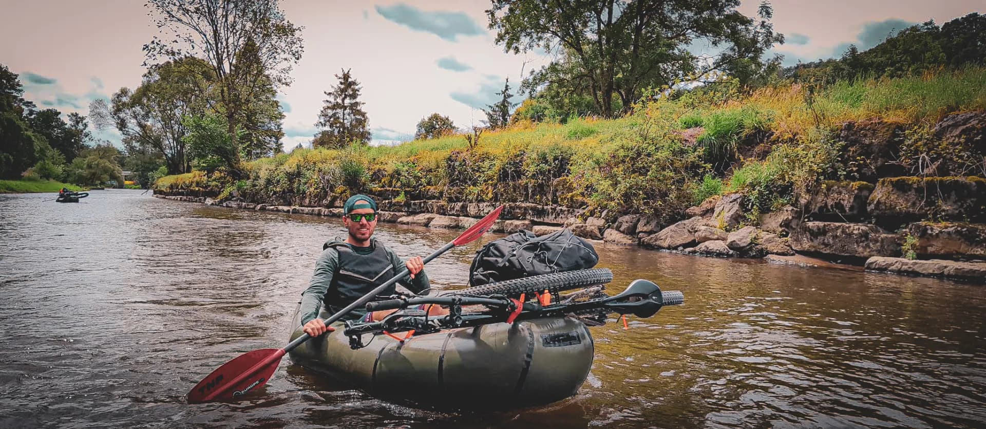A man paddling a small inflatable boat on a calm river, surrounded by lush green countryside. He is wearing sportswear, sunglasses and a hat, while his red paddle is visible.
