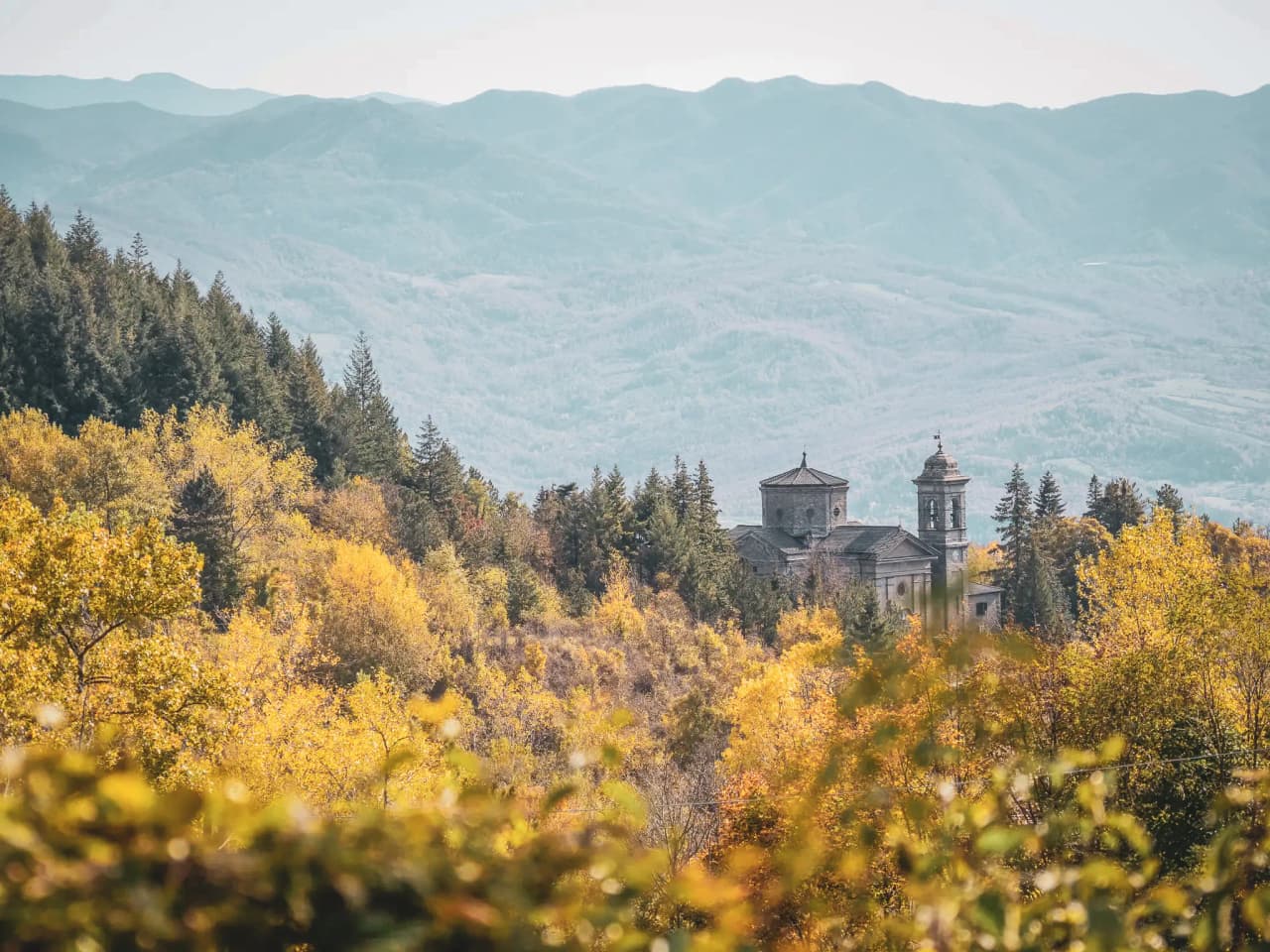 An autumnal landscape of trees with yellow and orange leaves, overlooked by mountains in the background. In the middle of the scene is an old religious-style building, probably a church or monastery, flanked by a