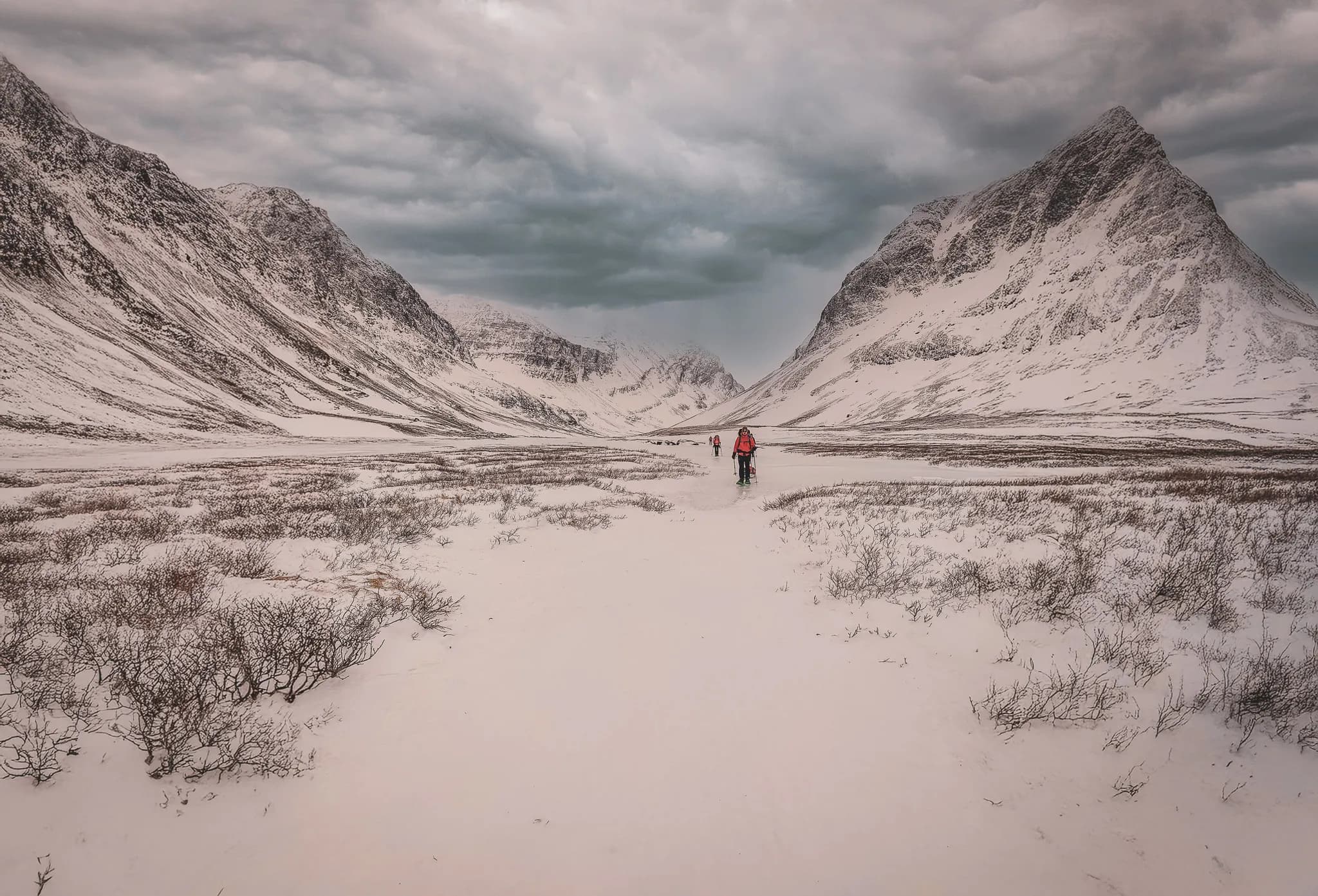 The mountainous landscape of Abisko in Swedish Lapland covered in snow, with steep peaks on either side. A group of people dressed in colourful outdoor clothing walk along a snowy path through a vast, wild valley.