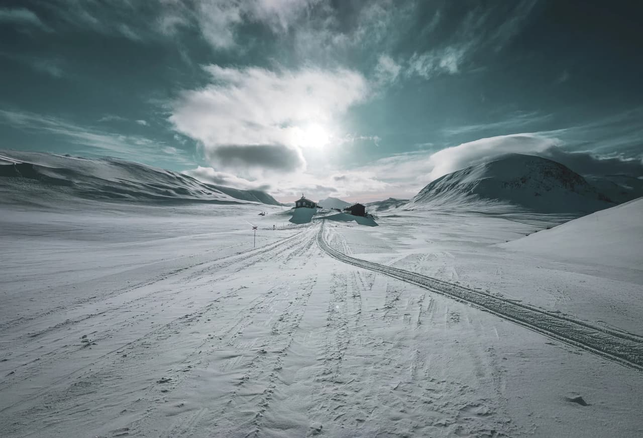 snowy landscape under a dramatic sky, with mountains and isolated cabins.