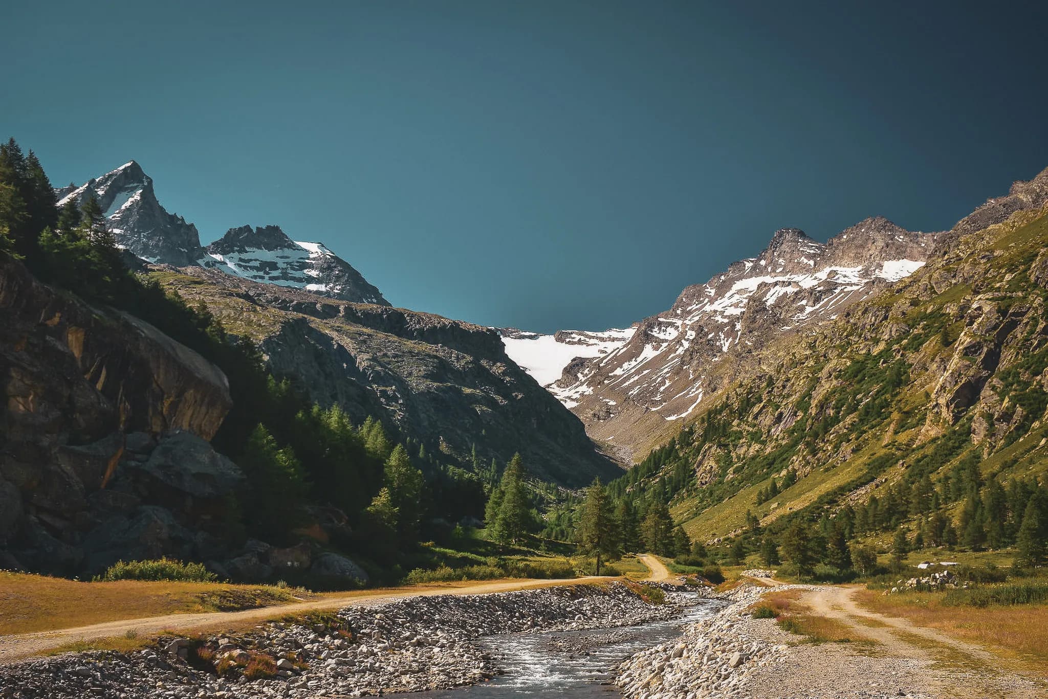 Een indrukwekkend berglandschap met besneeuwde toppen en rotsachtige hellingen. In het midden stroomt een kronkelende beek door een groene vallei, omringd door kleine keien.