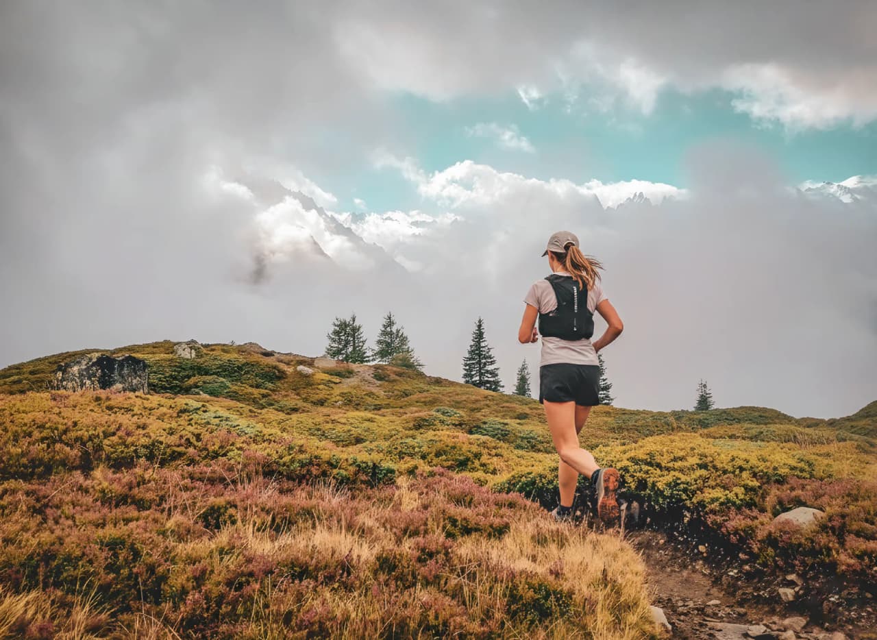 Femme courant sur un sentier alpin, entourée de paysages montagneux et de nuages impressionnants.