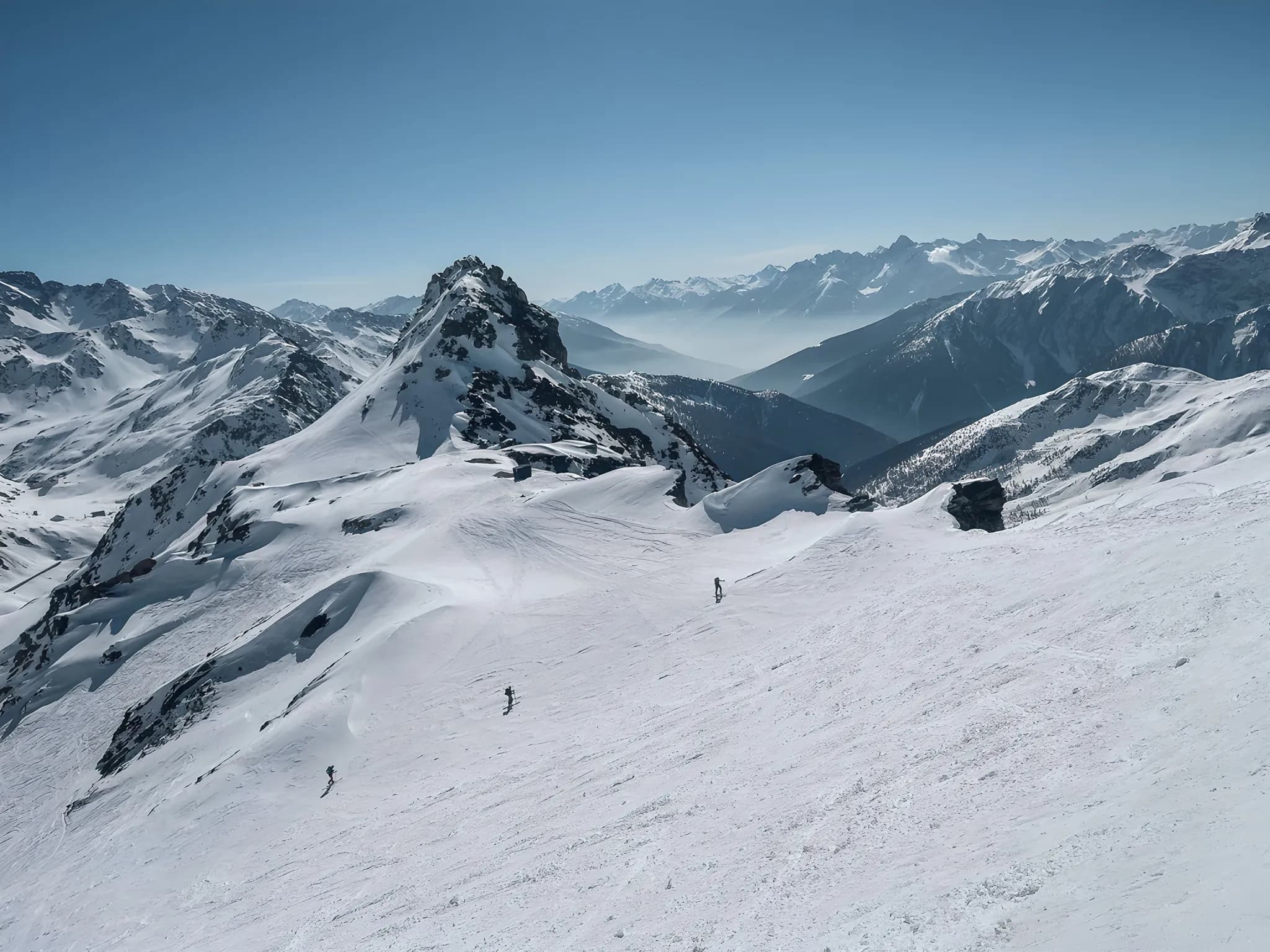 Snow-covered summit with skiers in an Alpine landscape, brilliant blue skies and majestic mountains.