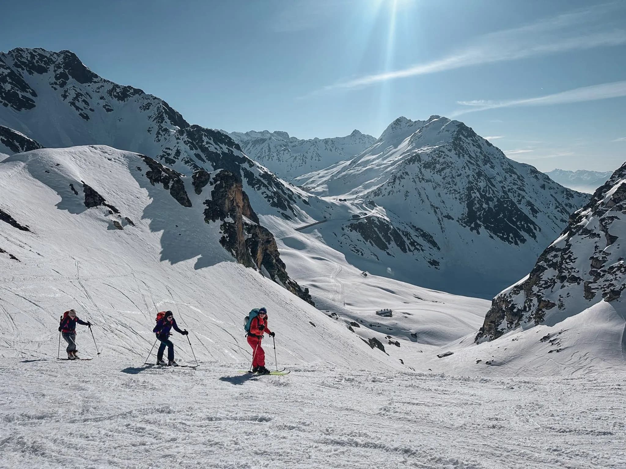 Three skiers explore a snow-covered alpine landscape under a brilliant blue sky, surrounded by majestic peaks.