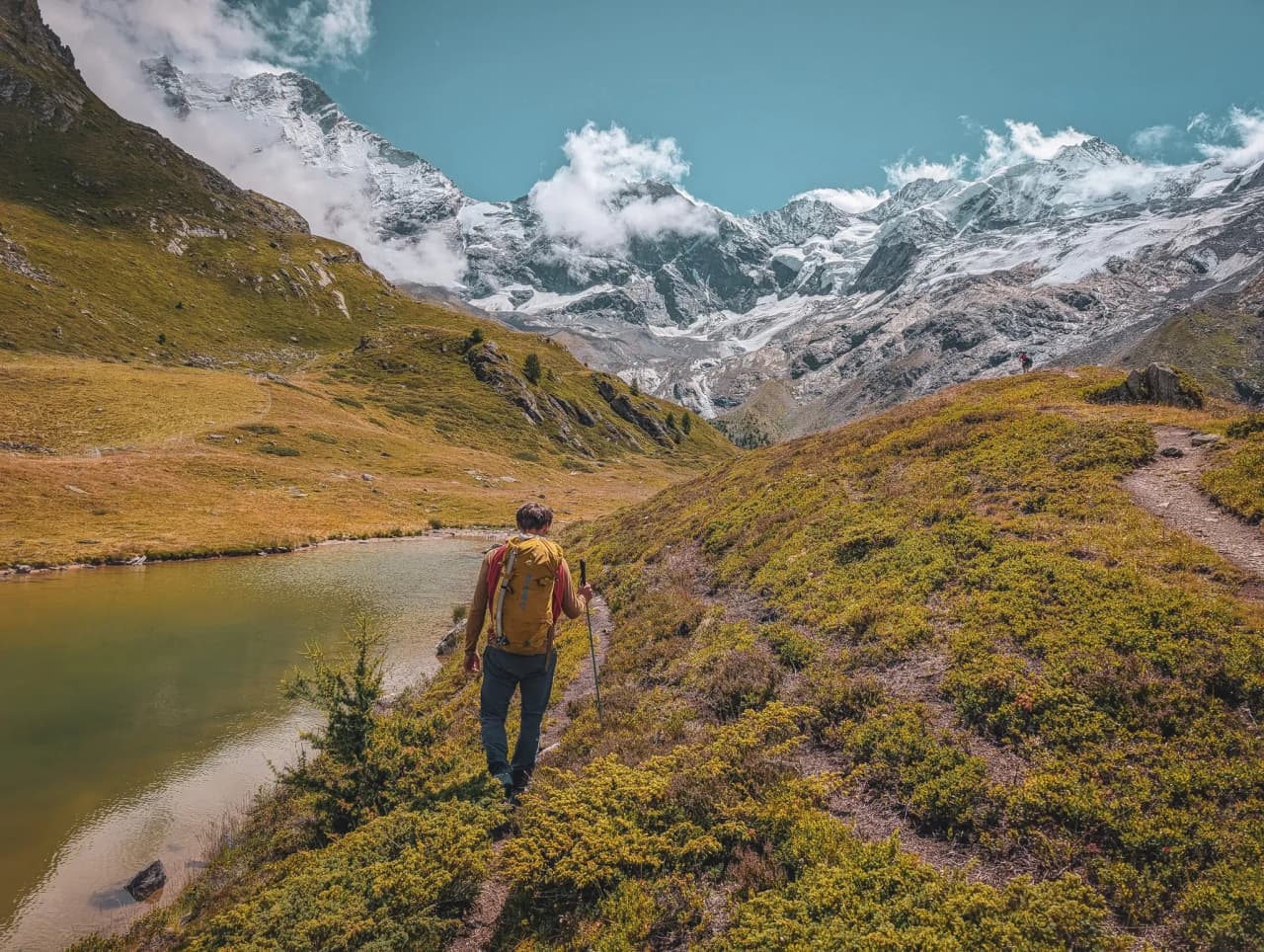 A solitary hiker hugs an Alpine lake, with the majestic snow-capped peaks in the background.