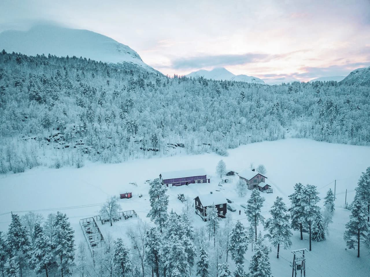 A snow-covered winter landscape, with a vast coniferous forest bordering a frozen lake. Wooden structures, including several houses and a farm building, are scattered across the landscape. The trees are rec