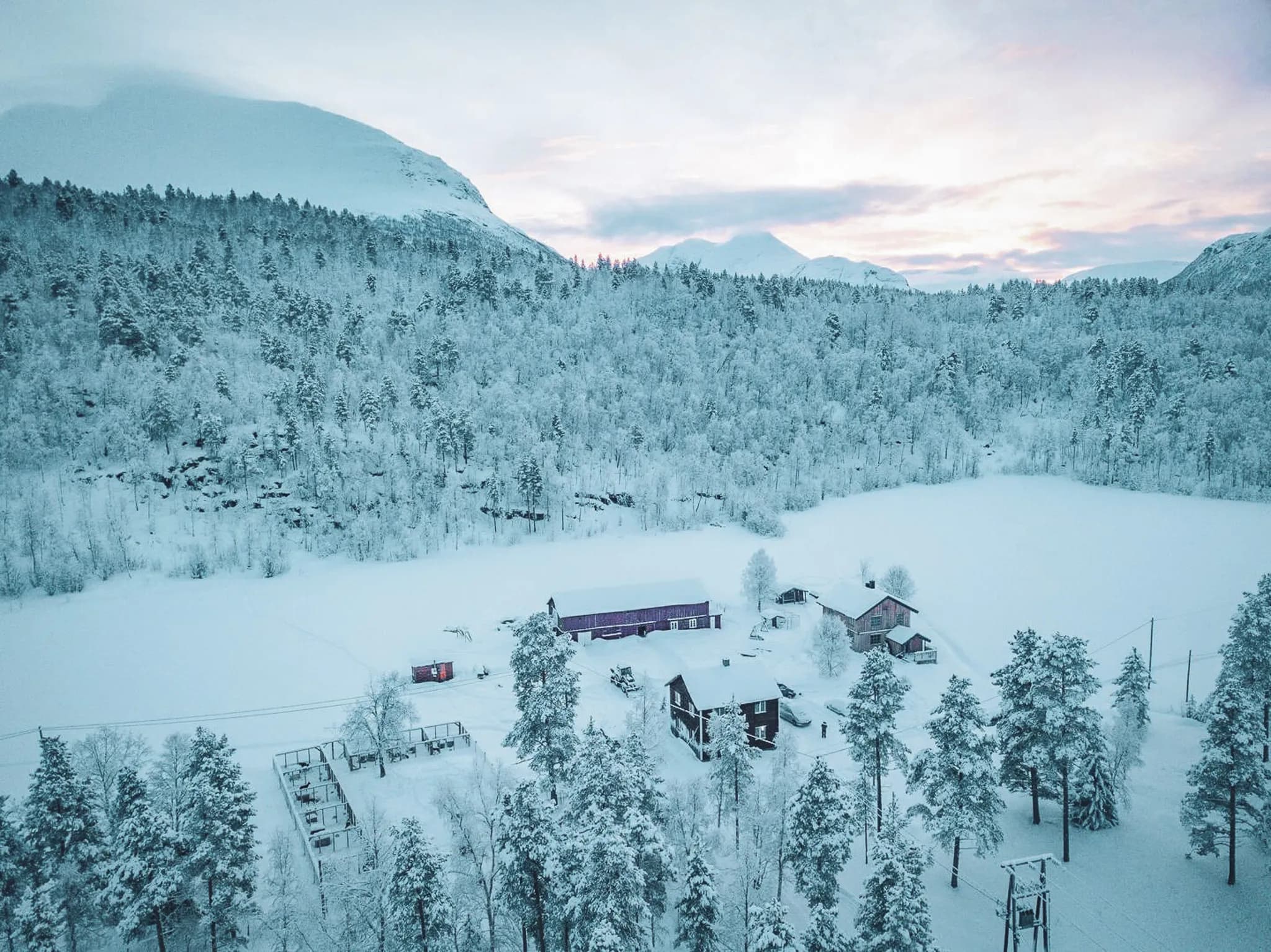 Un paysage hivernal couvert de neige, avec une vaste forêt de conifères bordant un lac gelé. Des structures en bois, dont plusieurs maisons et un bâtiment de ferme, sont éparpillées dans le paysage. Les arbres sont rec