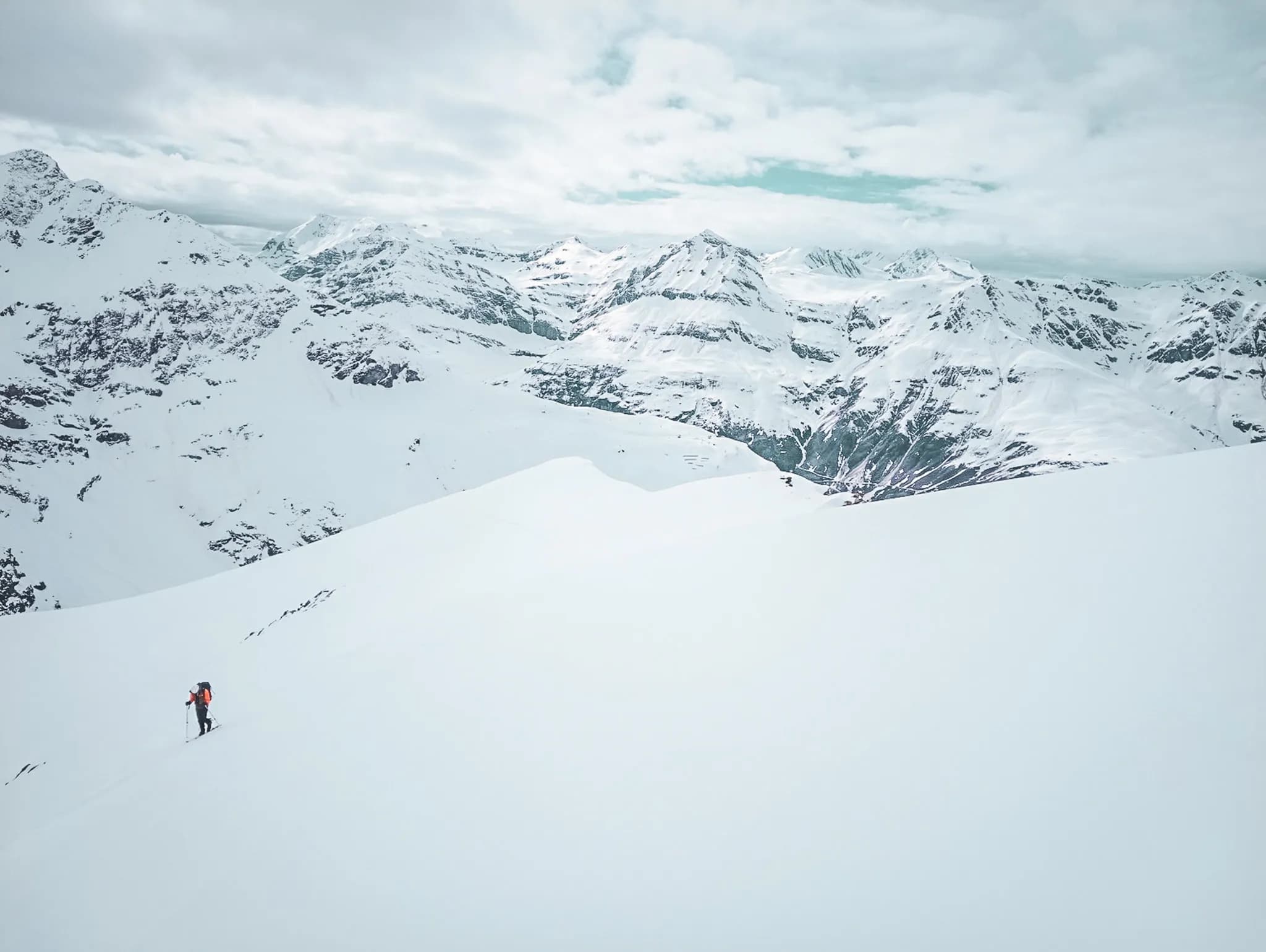 A moving skier explores a vast snow-covered landscape, surrounded by majestic snow-capped mountains. The sky is cloudy, creating an atmosphere of tranquillity, while the peaks of the mountains
