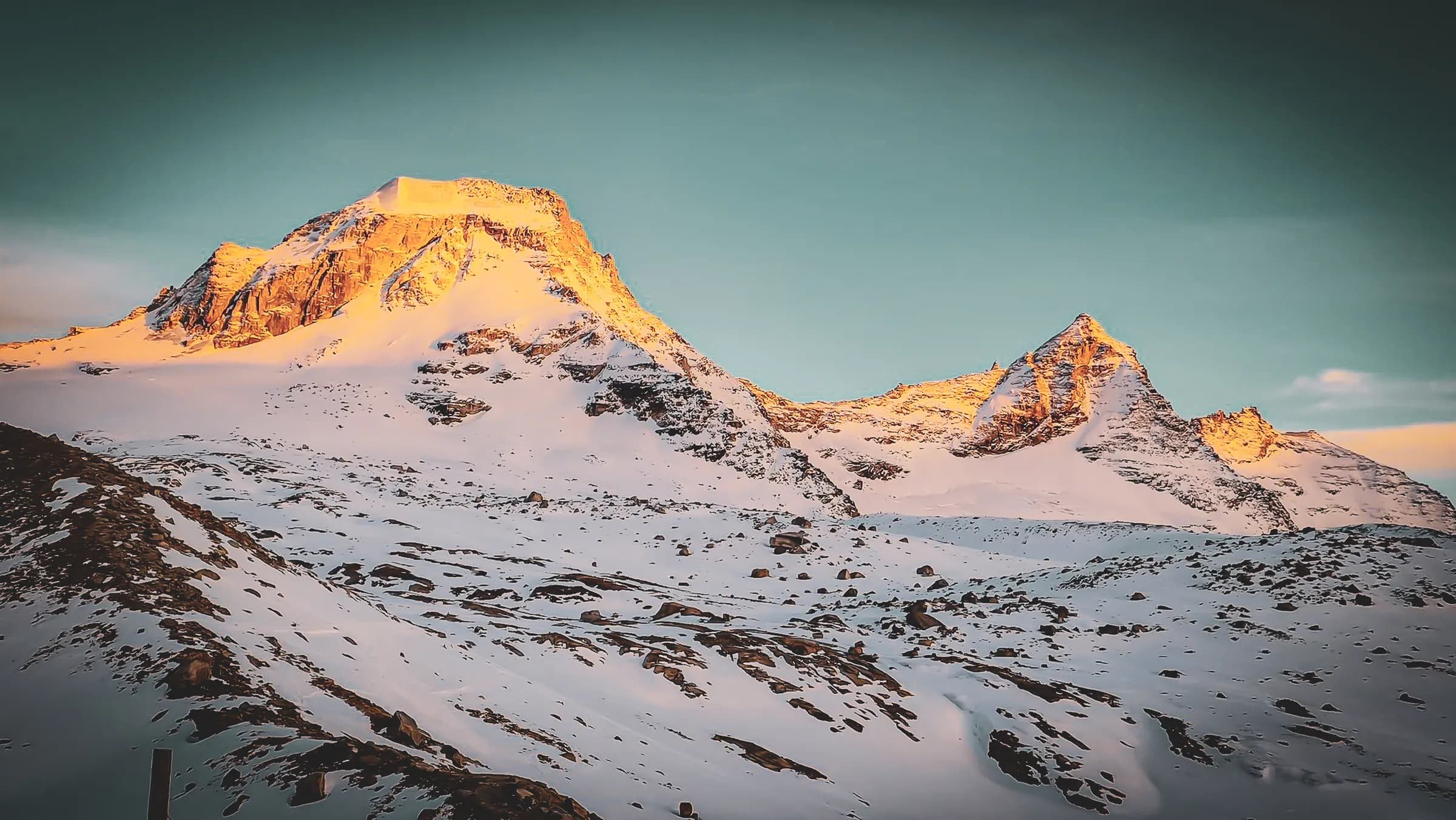 A majestic mountain landscape, where snow-capped peaks rise beneath a clear sky. Early morning sunlight illuminates the crest of one of the mountains, making the hardy snow bright and