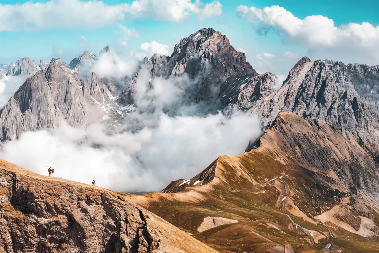 Two hikers at the top of a ridge, facing majestic, cloud-covered mountains.