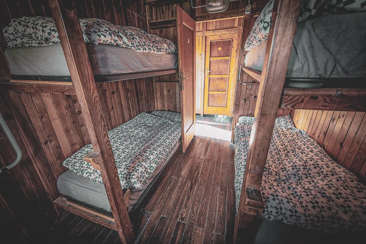 The warm interior of a wooden Mountain hut, with cosy bunk beds and an open door to adventure.