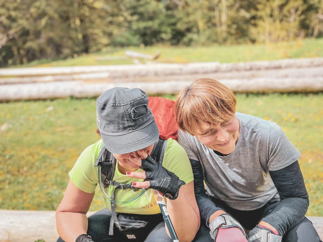 Two smiling women sitting on a section of tree, surrounded by greenery during a hike.