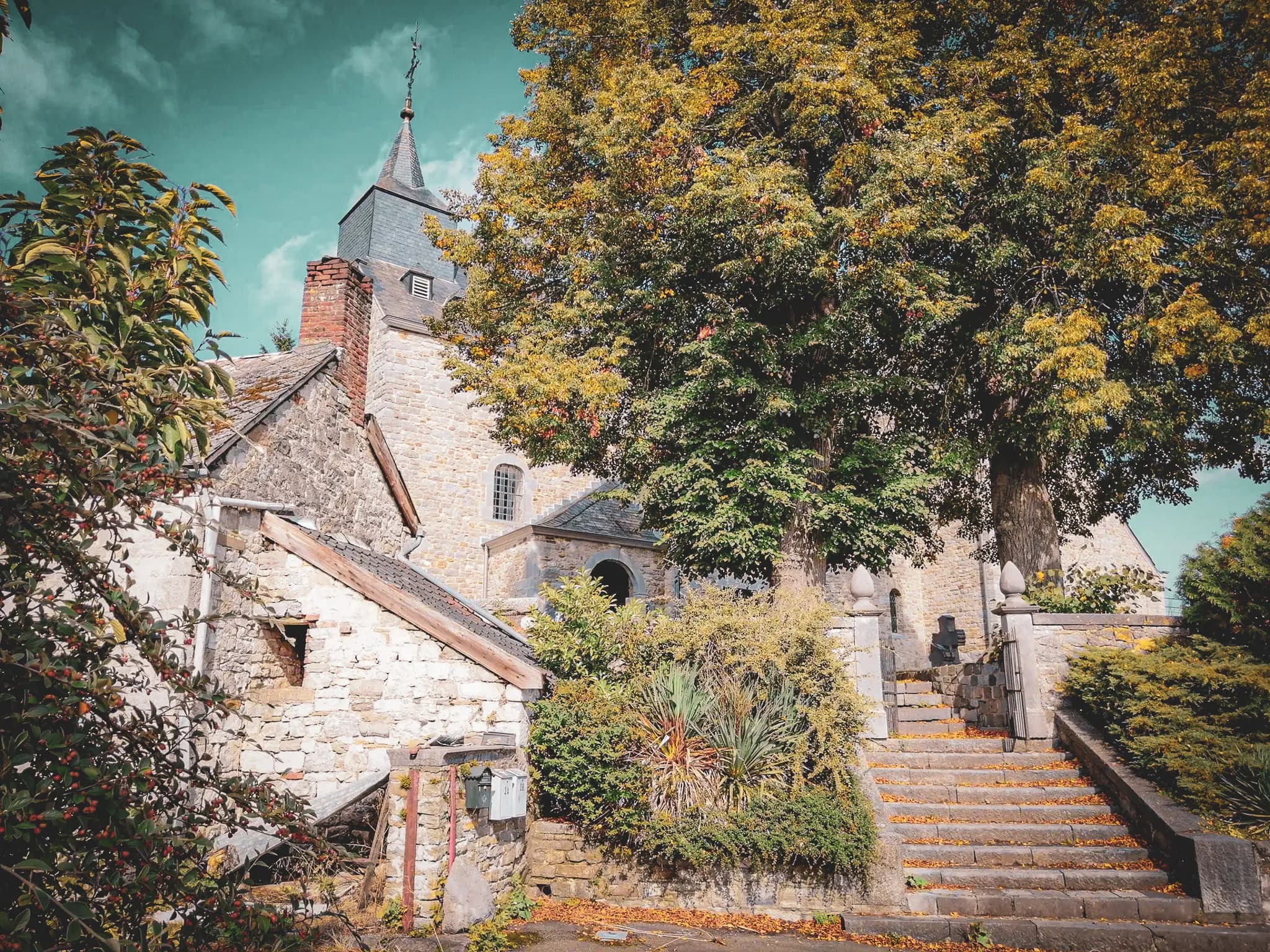 A picturesque village with a stone-walled church and pointed bell tower. Stone steps lead up to the church entrance, lined with fallen tree leaves. A large tree