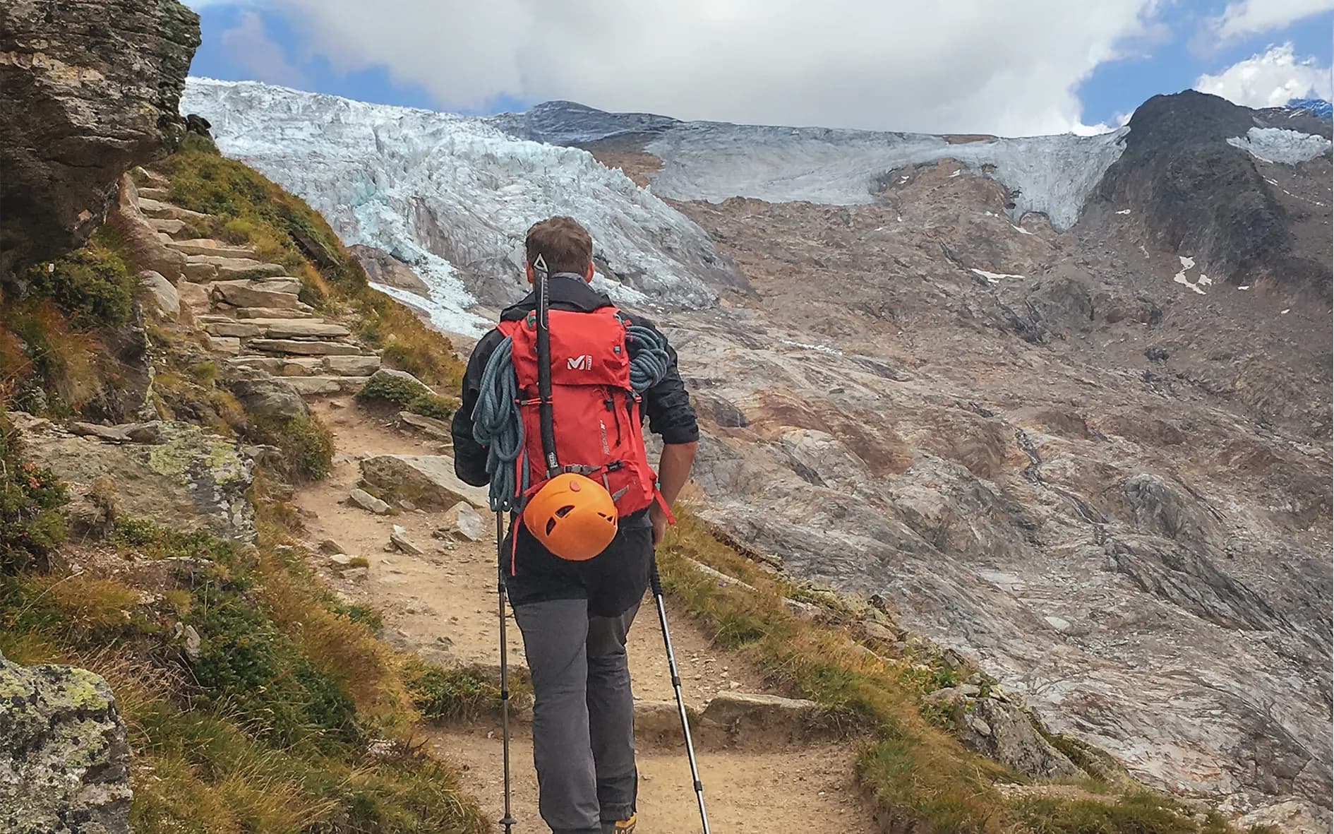 A hiker heads towards a majestic glacier, wearing a red rucksack and an orange helmet. He follows a steep path, lined with rocks and green grass, with steps in