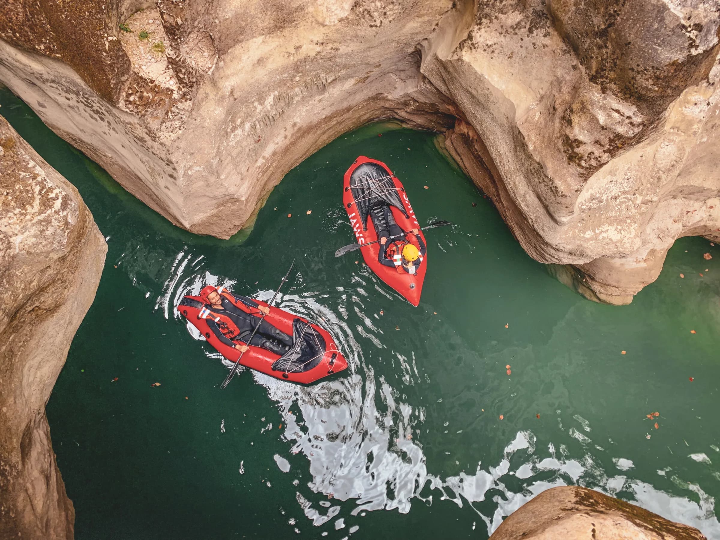 Two kayakers navigate a narrow canyon with rocky walls overlooking blue-green water. The kayaks are red and one of the paddlers is wearing a helmet and wetsuit, while the other is wearing a wetsuit.
