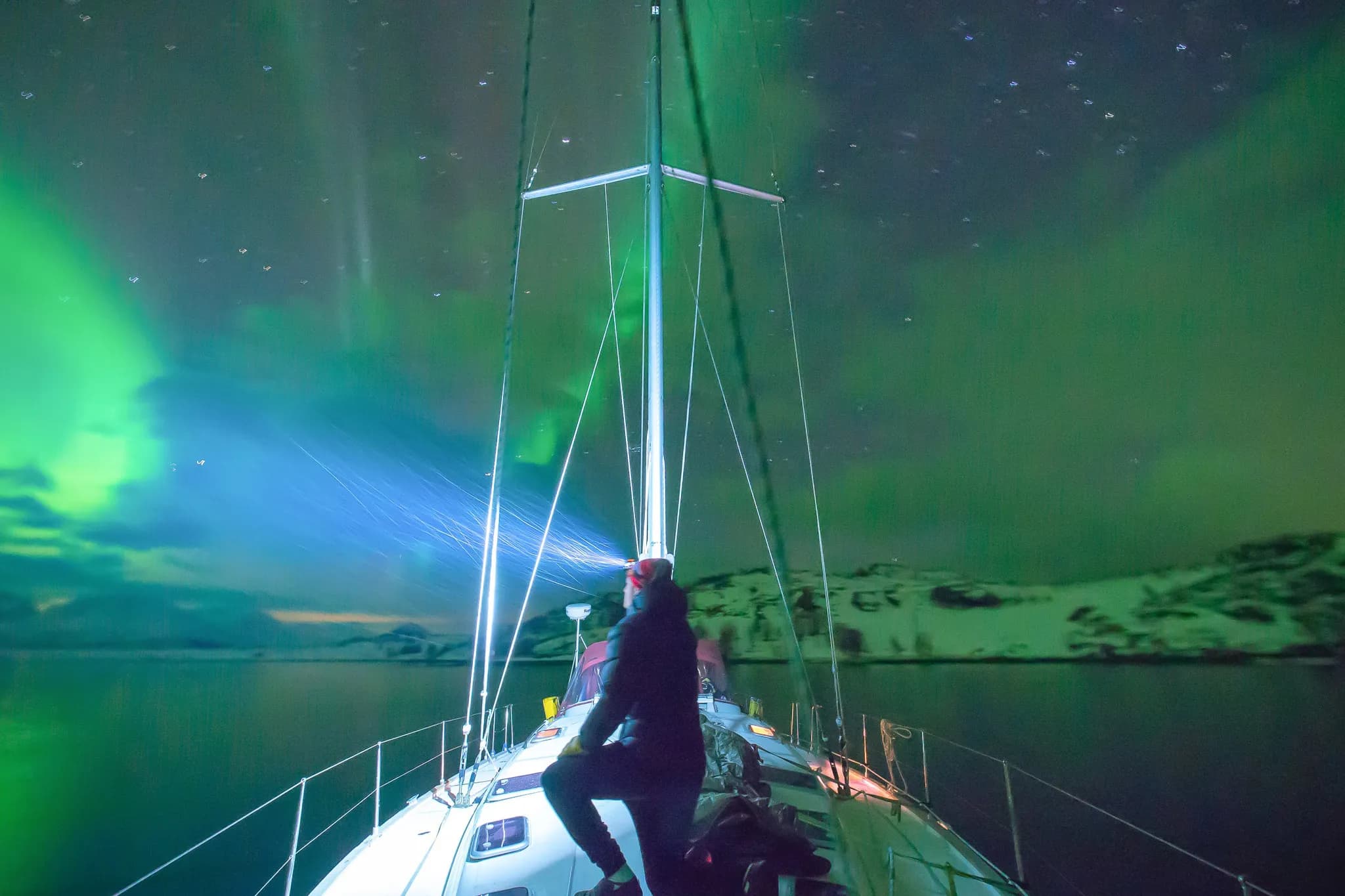 Un voilier navigue à travers des eaux calmes sous un ciel illuminé par des aurores boréales vertes et bleues. À l'avant du bateau, une personne, vêtue d'un manteau sombre et portant une lampe