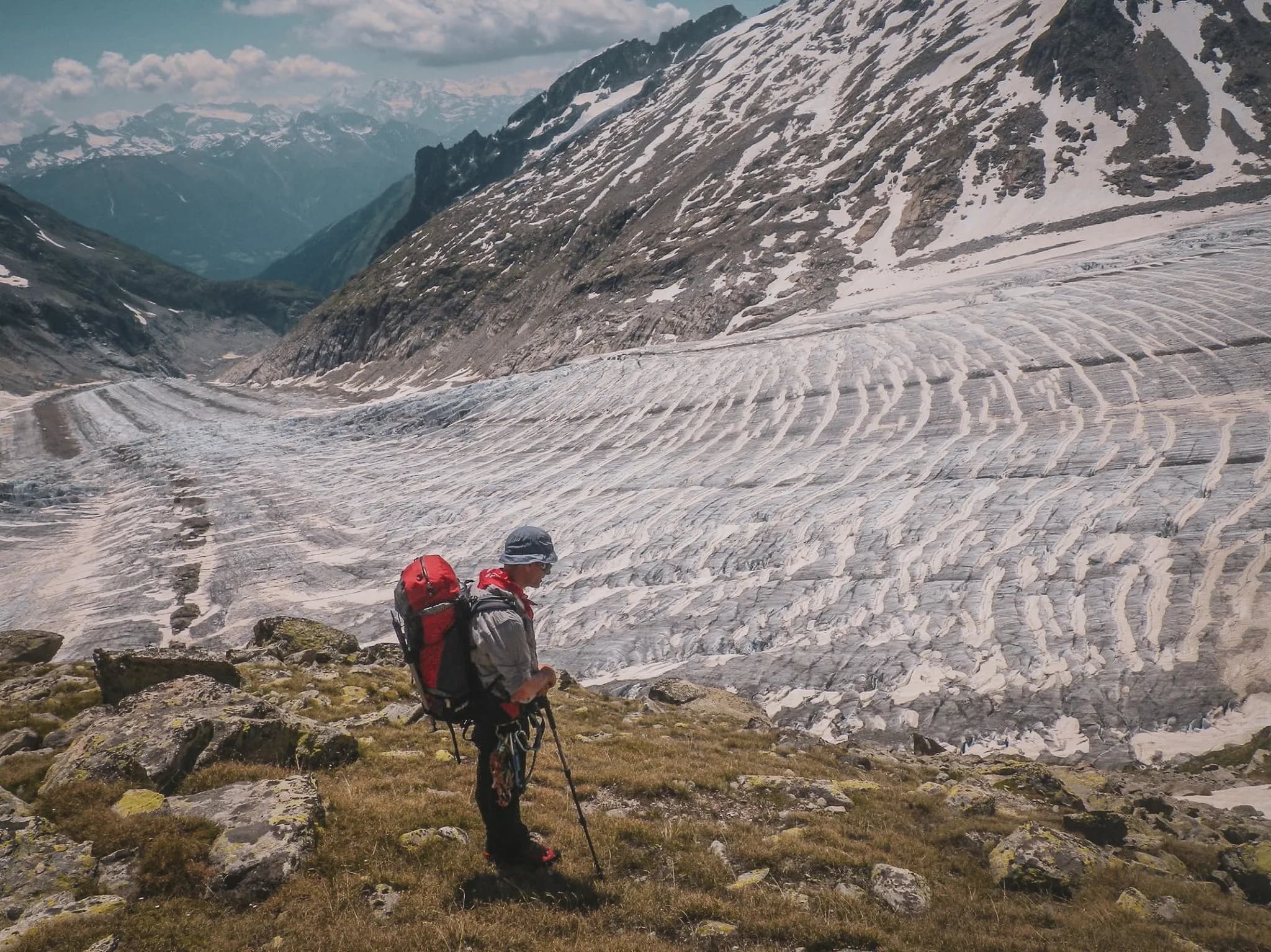 A climber stands on rocky terrain overlooking the largest glacier in Europe. The undulating ice stretches as far as the eye can see, surrounded by majestic mountains.
