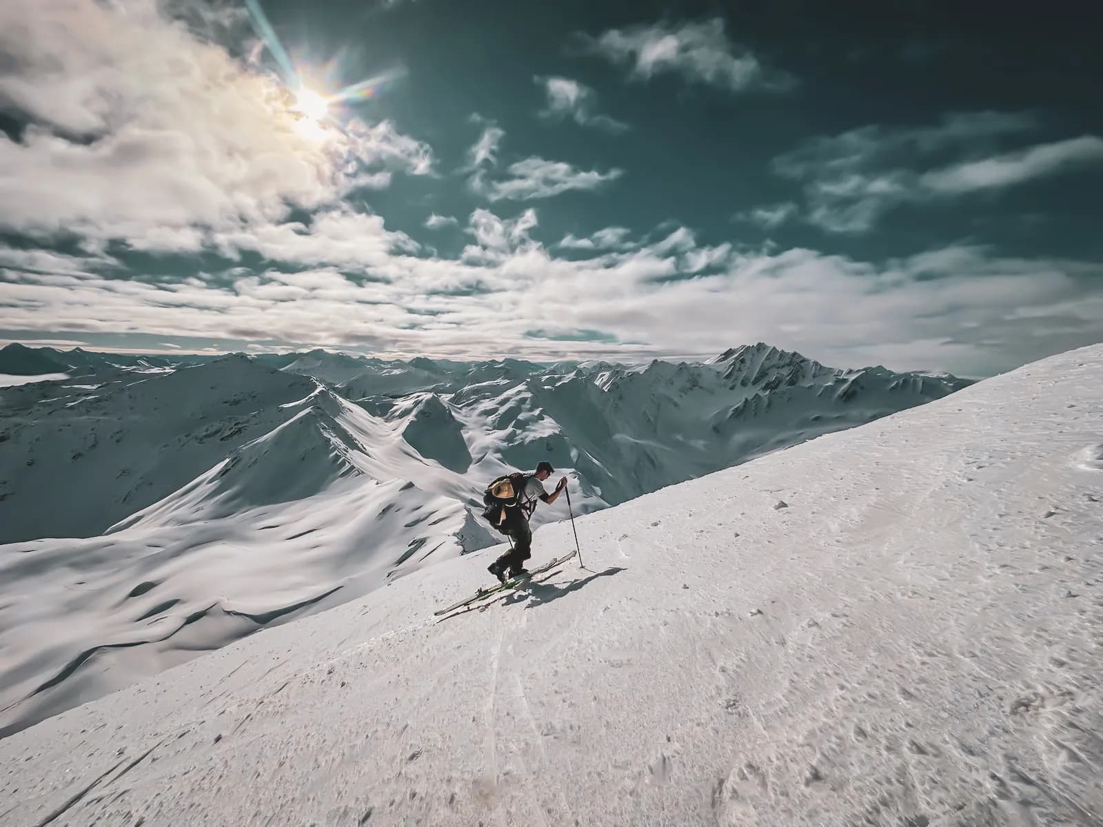 A skier climbing a snow-covered mountainside, surrounded by majestic peaks. The sky is partly cloudy, with the sun shining brightly. The immaculate snow