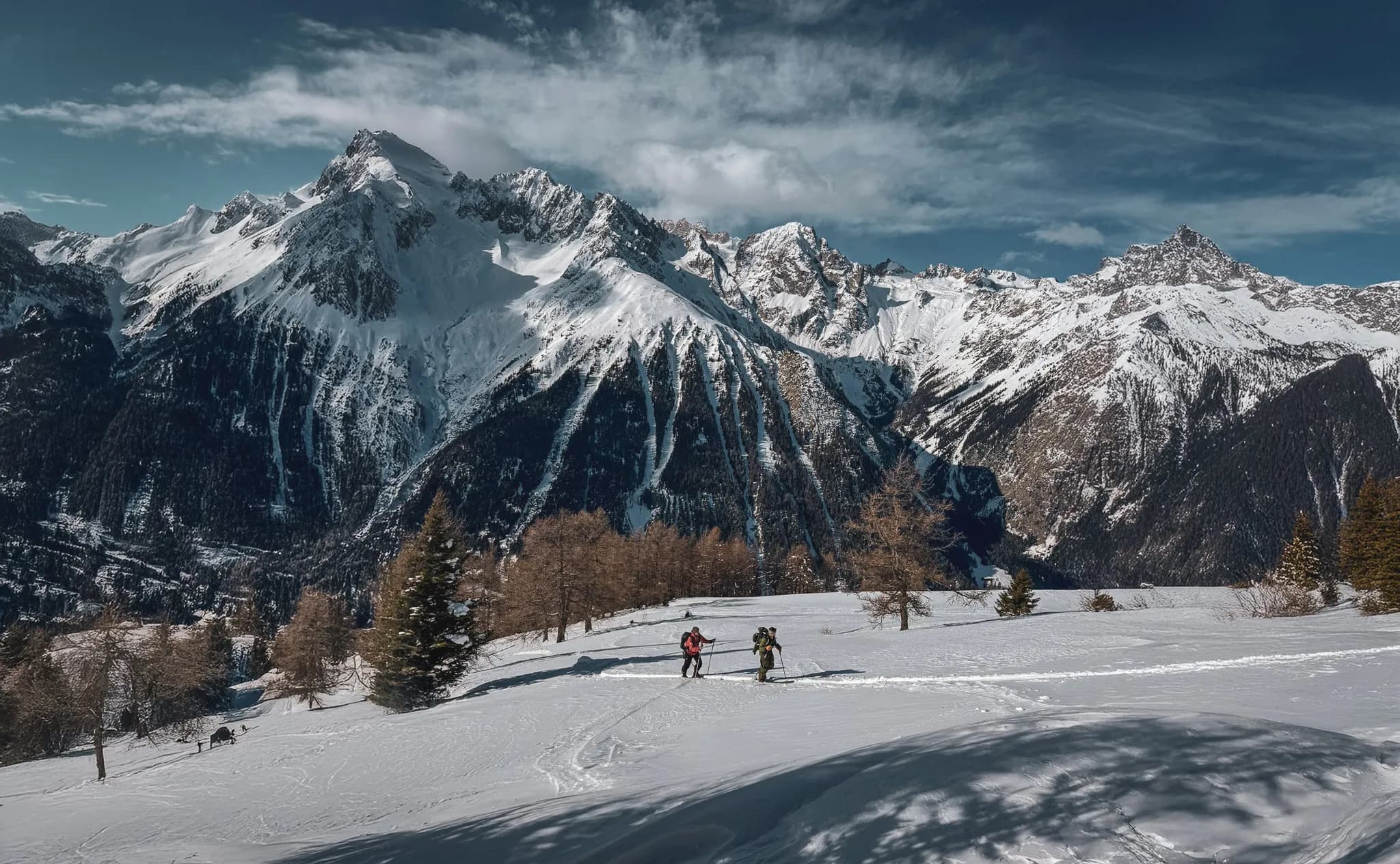 A spectacular Alpine landscape with majestic snow-capped mountains rising up under a slightly cloudy sky. In the foreground, two skiers are making their way across a fresh expanse of snow, surrounded by