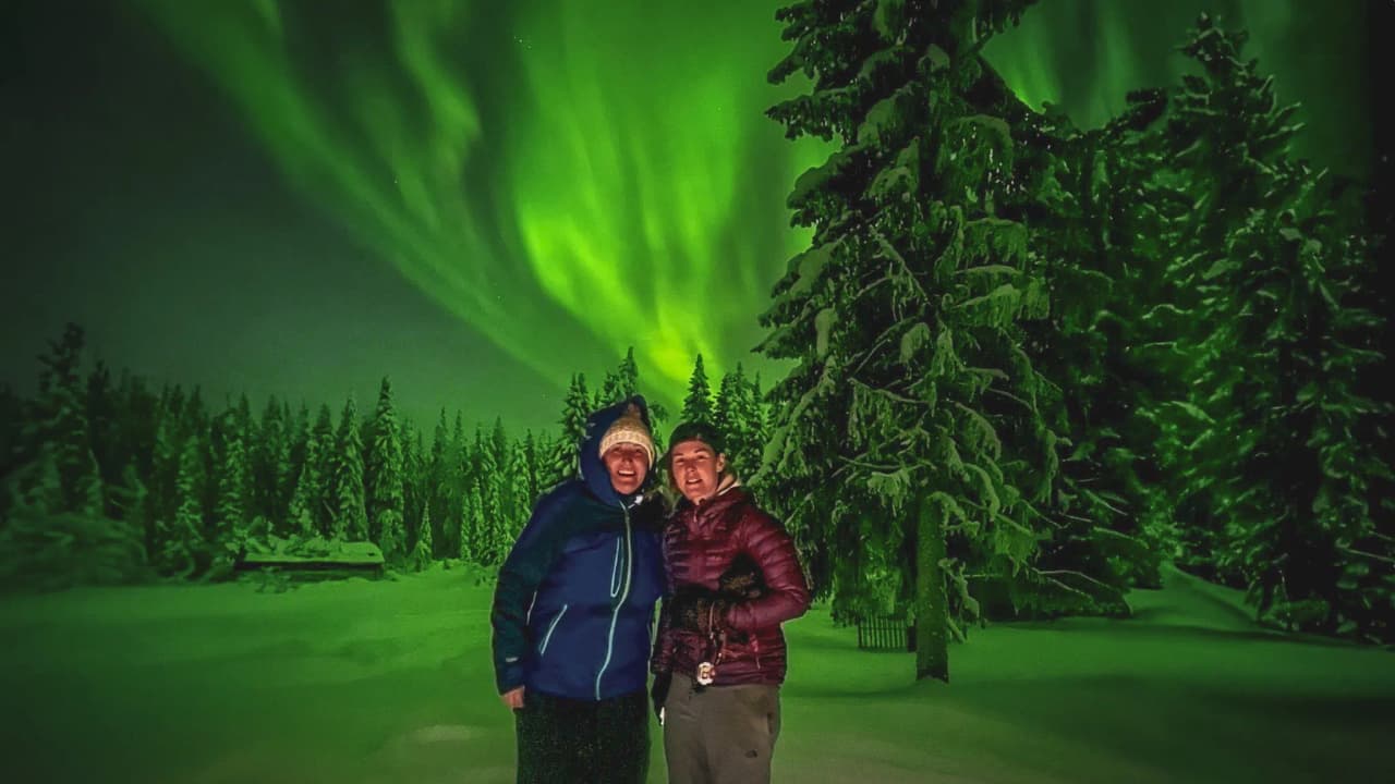 Two smiling people under the northern lights, surrounded by a snow-covered forest in Lapland.