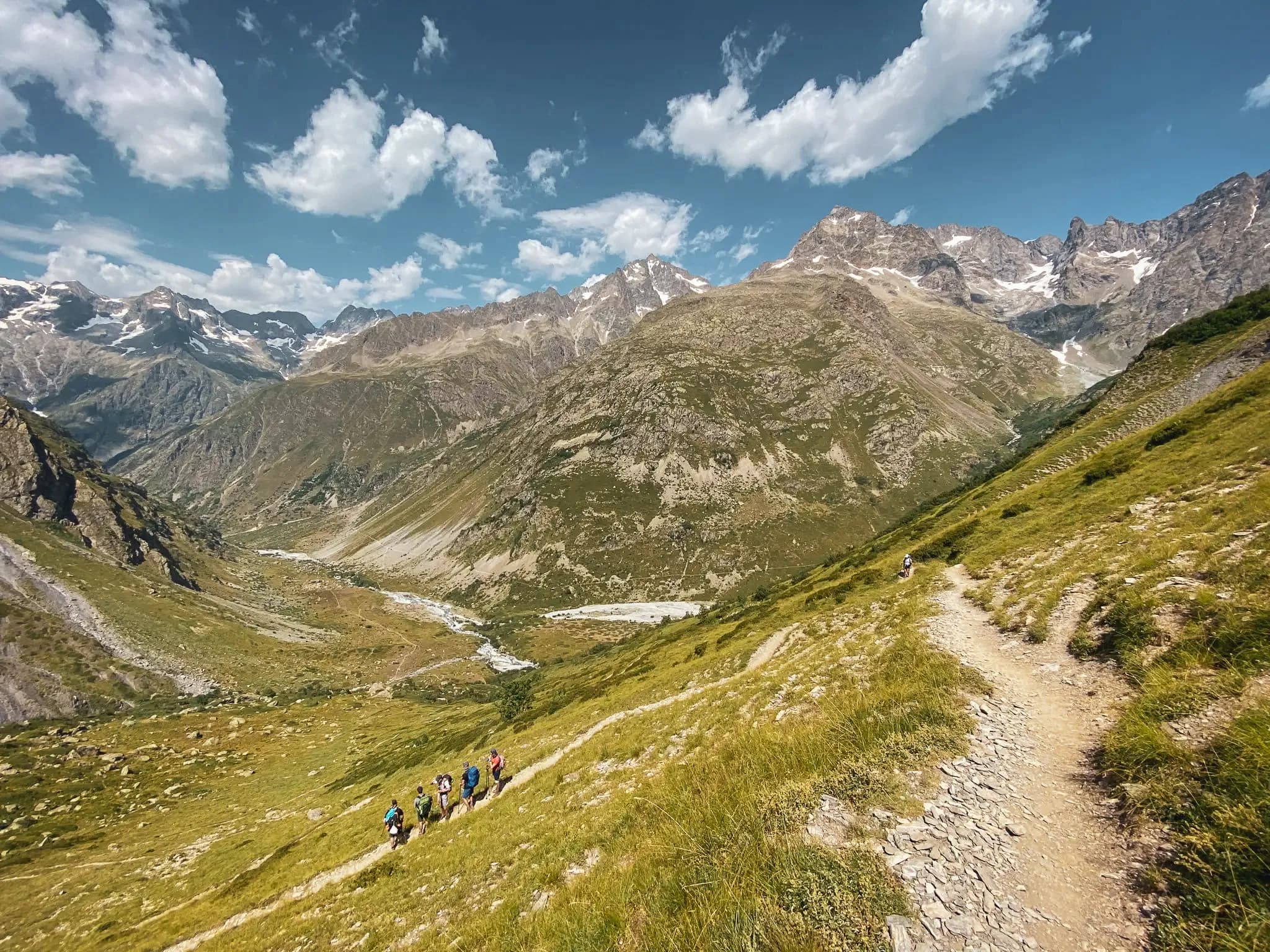 Wandelaars kronkelen over een alpenpad, omringd door majestueuze bergen en blauwe luchten.