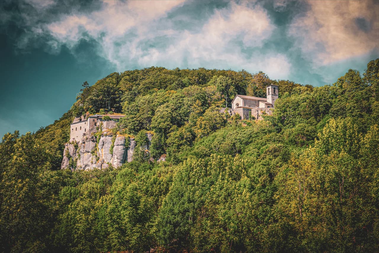 The monastery of Camaldoli is perched on a green hill, surrounded by dense woodland. The stone structure features typical architectural elements, such as a bell tower and arched windows.