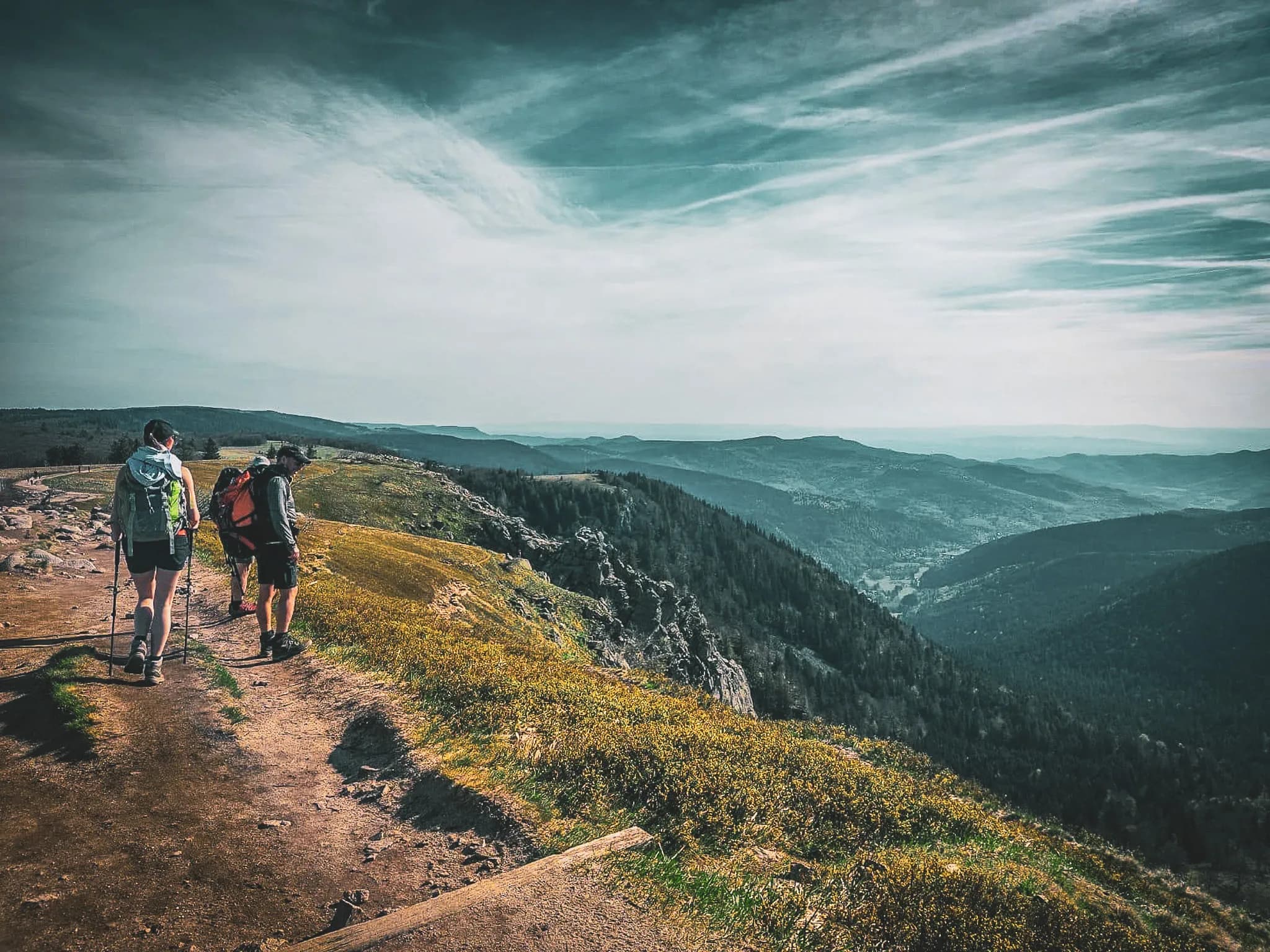 A mountain hiking trail, with two people walking side by side. The atmosphere is peaceful, with a vast view of the valley stretching out in the background, surrounded by hills and