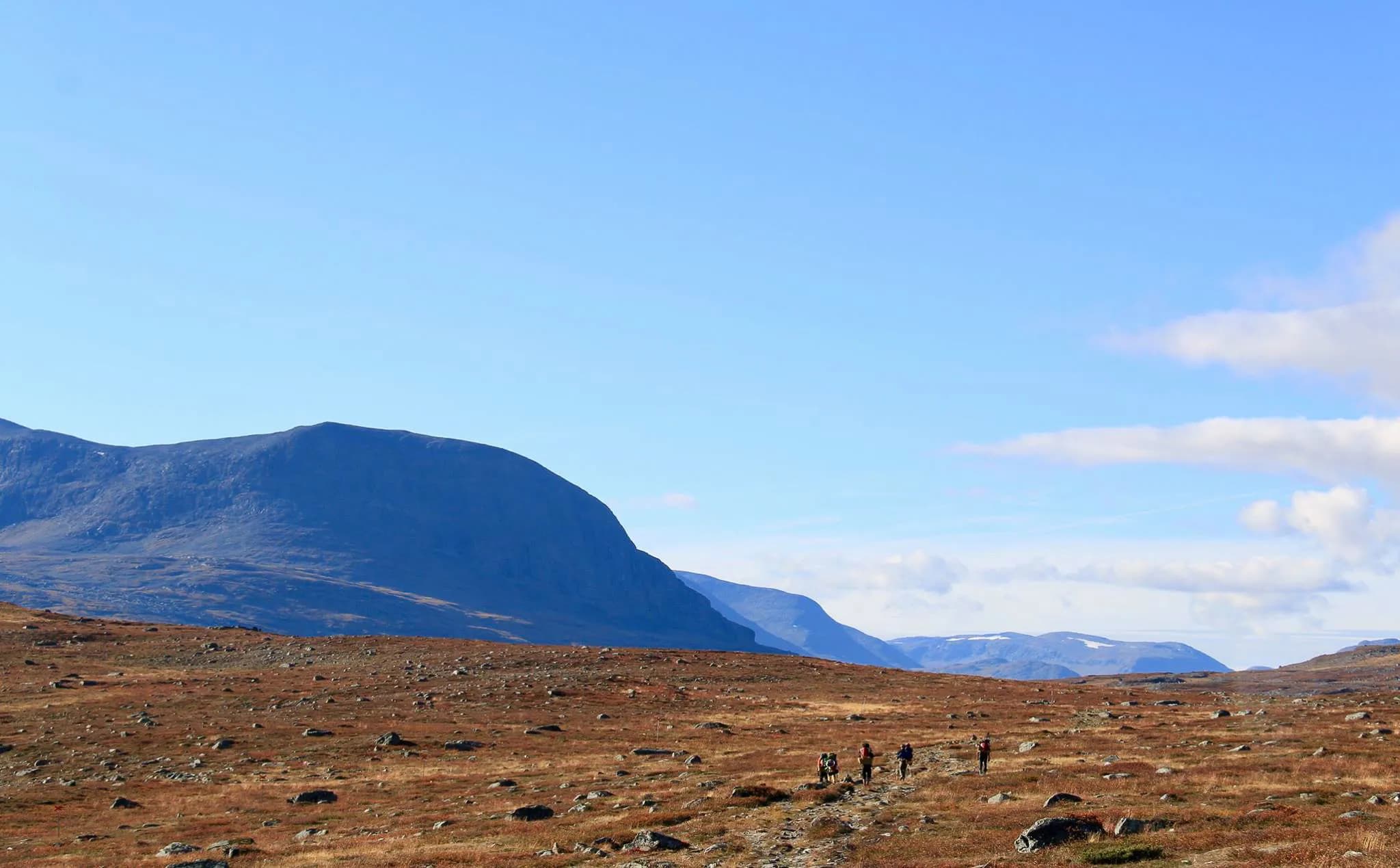 A mountainous landscape with hills and peaks in the background, under a clear blue sky dotted with a few white clouds. In the foreground, a vast expanse of arid, gently undulating terrain, dotted with