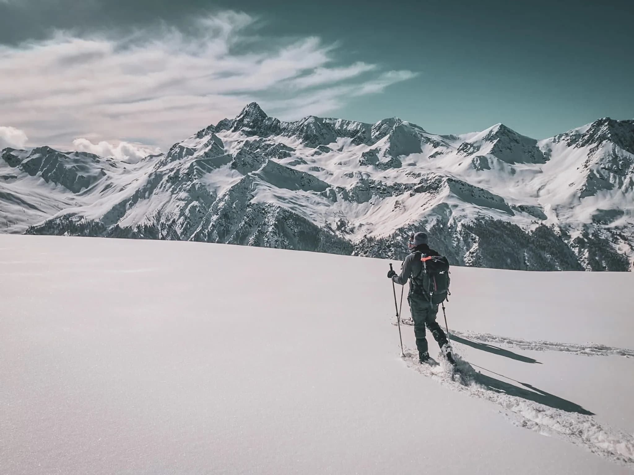 A lone skier on a vast field of immaculate snow, with majestic mountains in the background. The sky is clear, dotted with light clouds, and the day's light illuminates the snow.