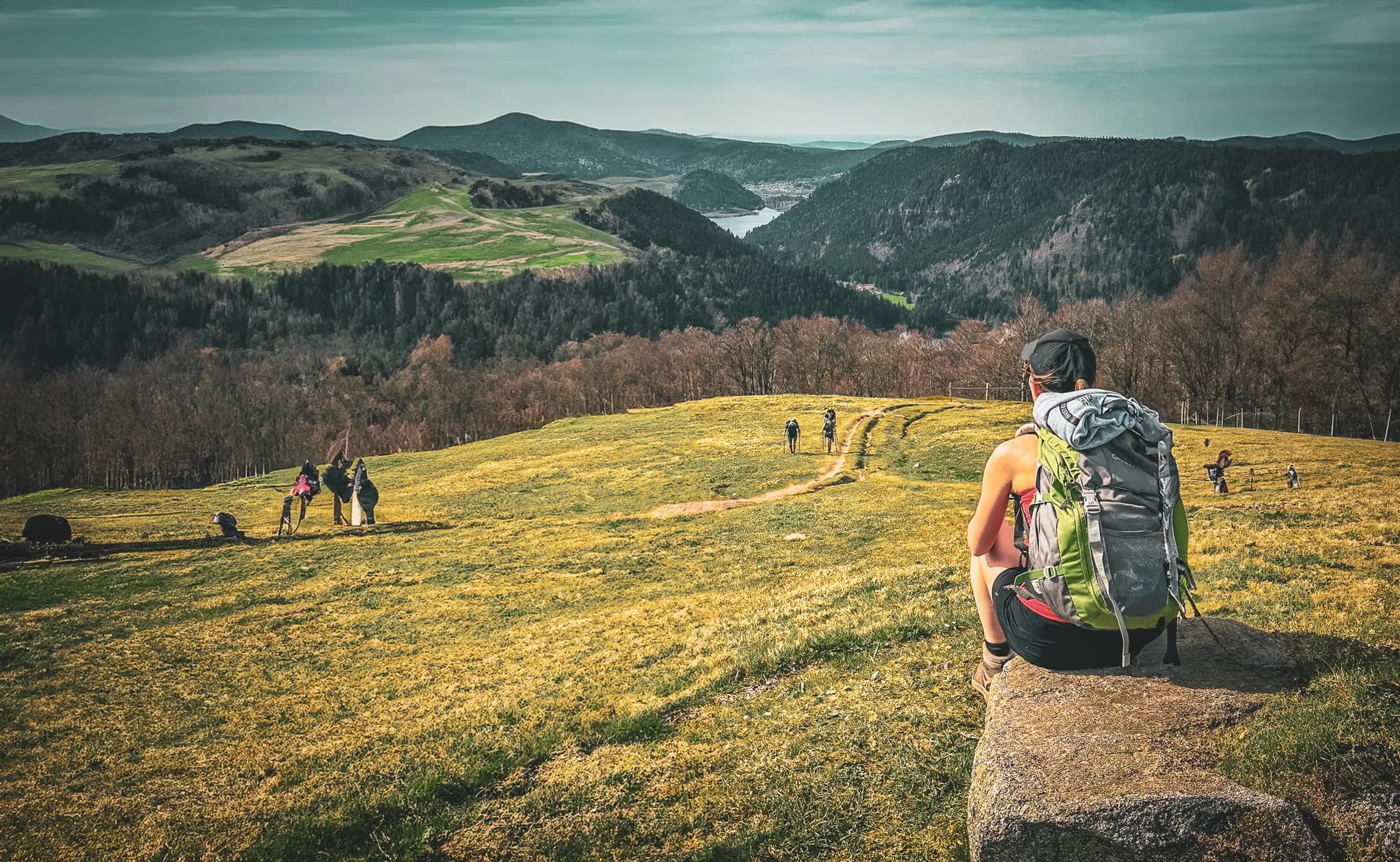 A hiker sitting on a rock, contemplating a landscape of green hills and mountains in the distance. The terrain is dotted with yellow grass and winding paths, with other hikers