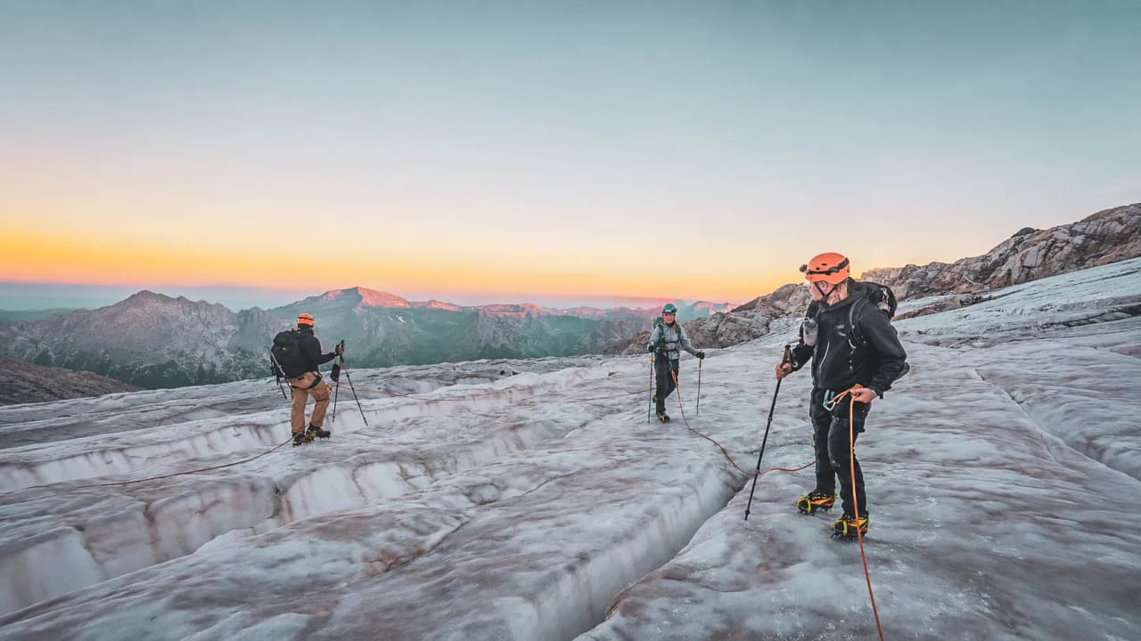 Groupe d'alpinistes sur un glacier au lever du soleil, avec des sommets majestueux en arrière-plan.