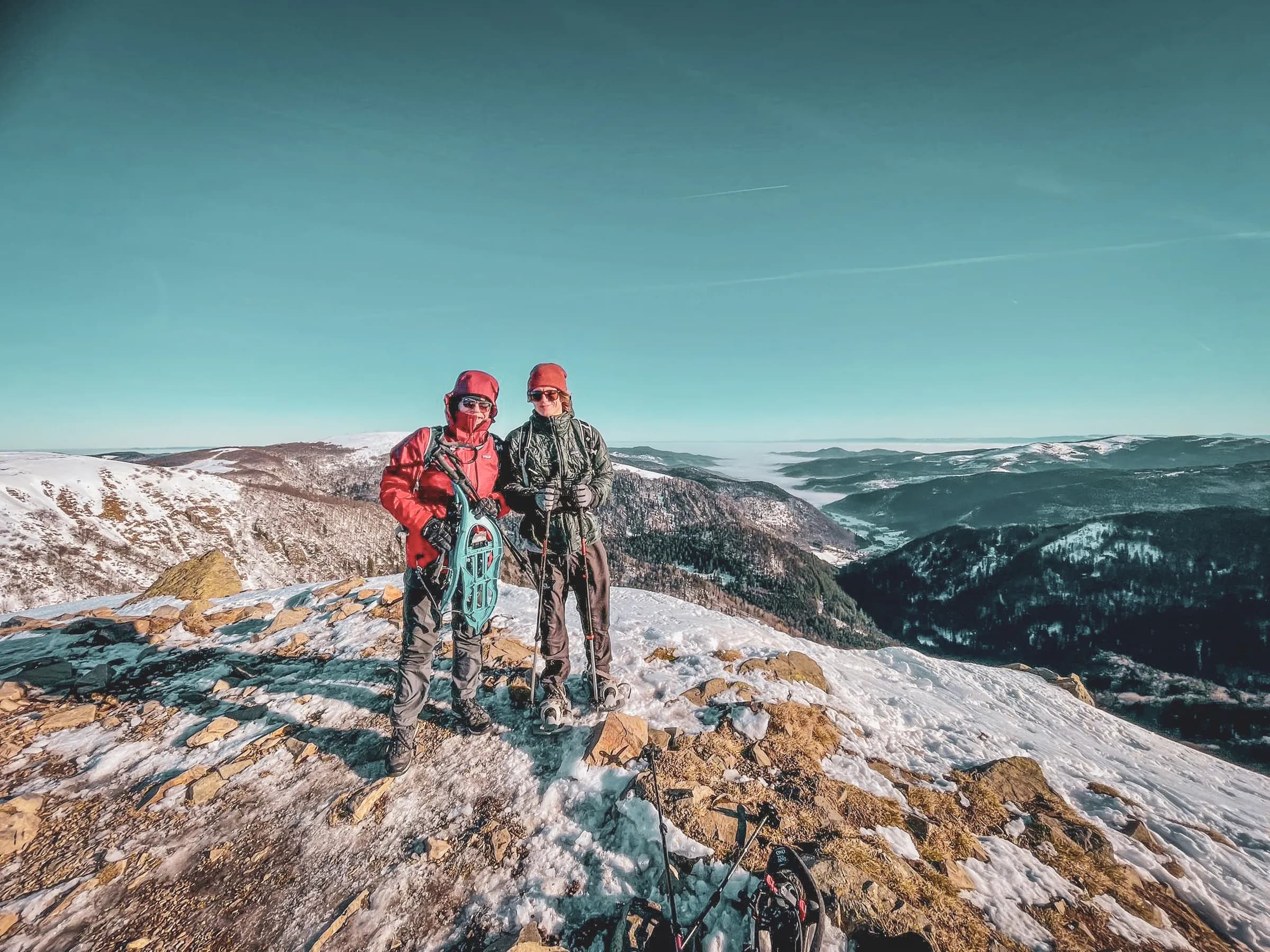 Twee mensen poseren op de top van een besneeuwde berg, gekleed in outdoorkleding die geschikt is voor de kou, waaronder een rode jas en muts. Ze houden sneeuwschoenen en een stok vast en tonen een