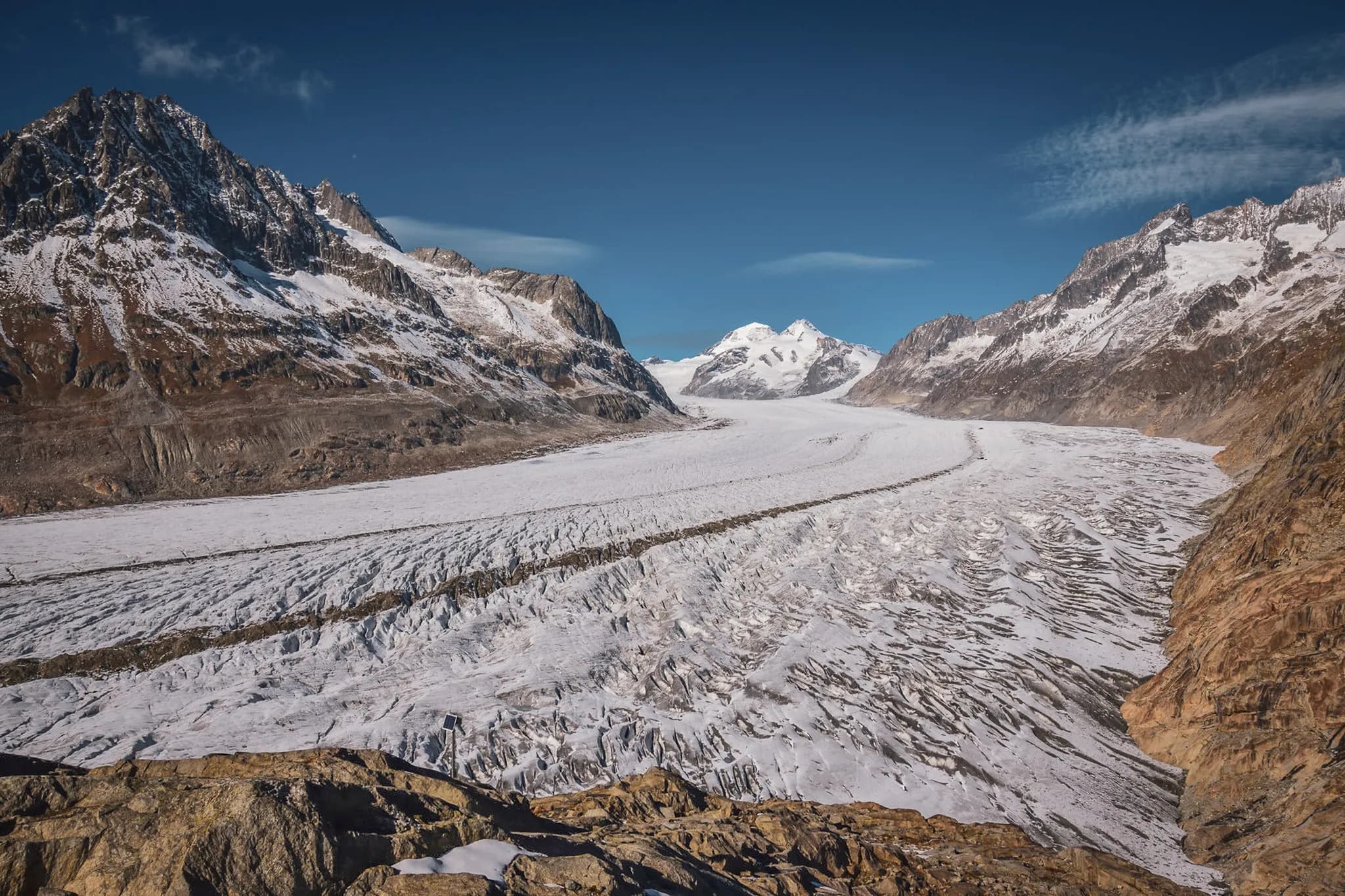 A vast glacier stretches between mountain peaks, with rocky, snow-covered slopes. The surface of the glacier is marked by crevasses and striations, while the sky is clear.