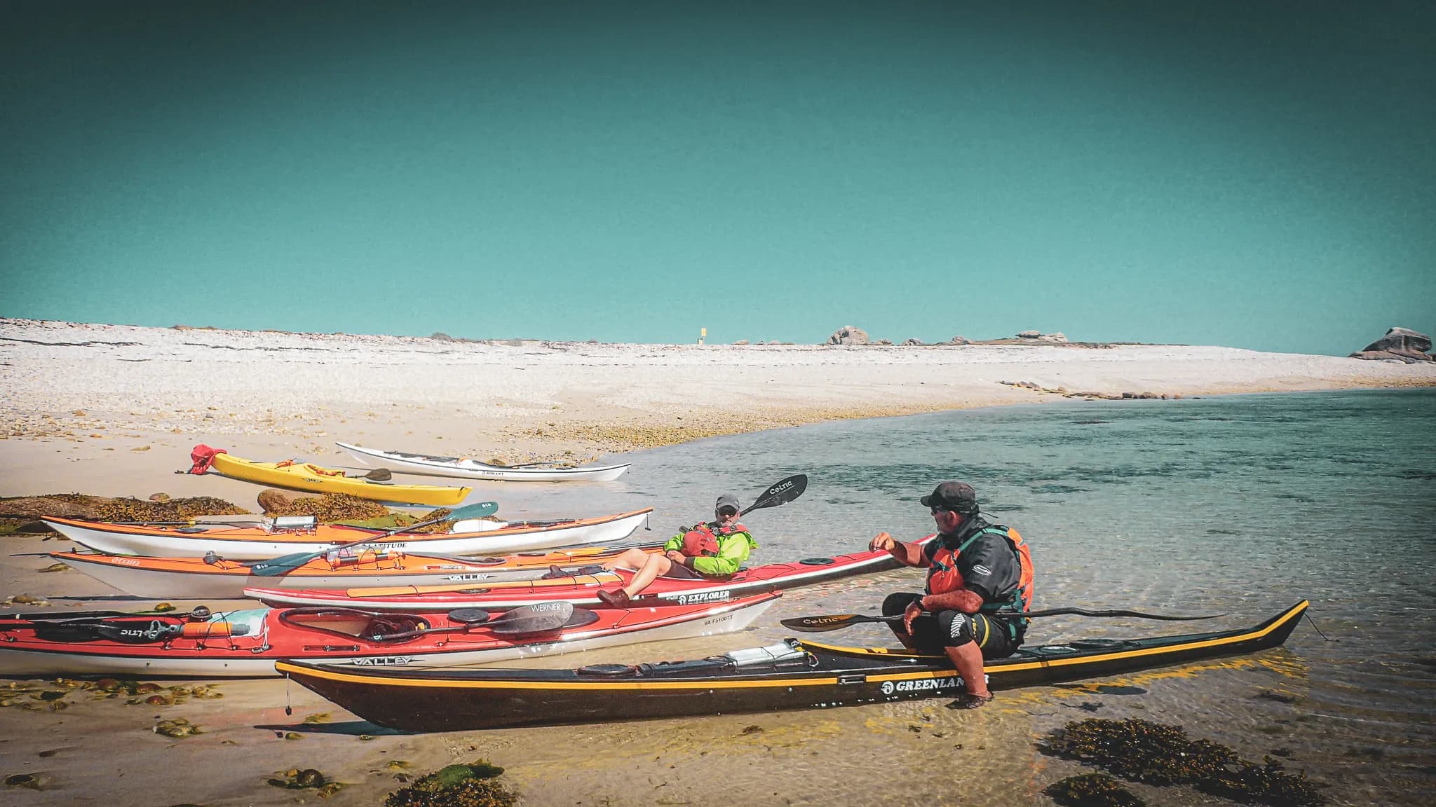 A group of kayaks of different colours are resting on a sandy beach near calm water. Two people, one lying on a red kayak and the other sitting on a black kayak, are chatting quietly.