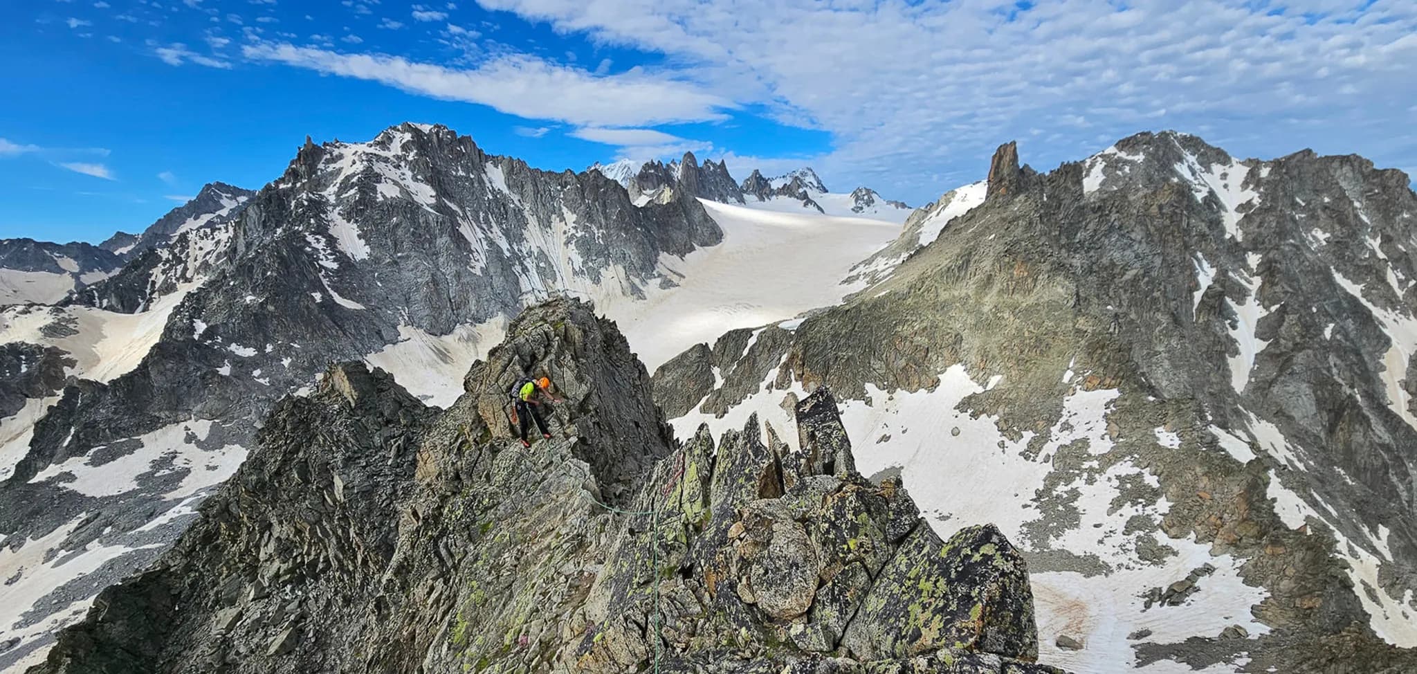 A majestic mountain landscape emerges, with towering rocky peaks and sparkling glaciers. A mountaineer wearing an orange helmet and harness climbs up a steep ridge, surrounded by a rocky outcrop.
