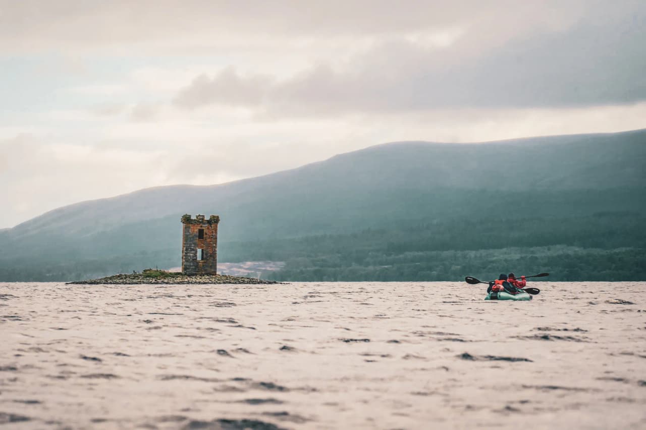 Zeil met een packraft op een vredig loch aan de voet van de Highlands, met een schilderachtige toren.