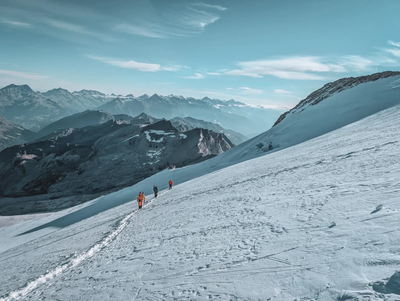 Climbers make their way across a glacier, surrounded by majestic mountains under a blue sky.