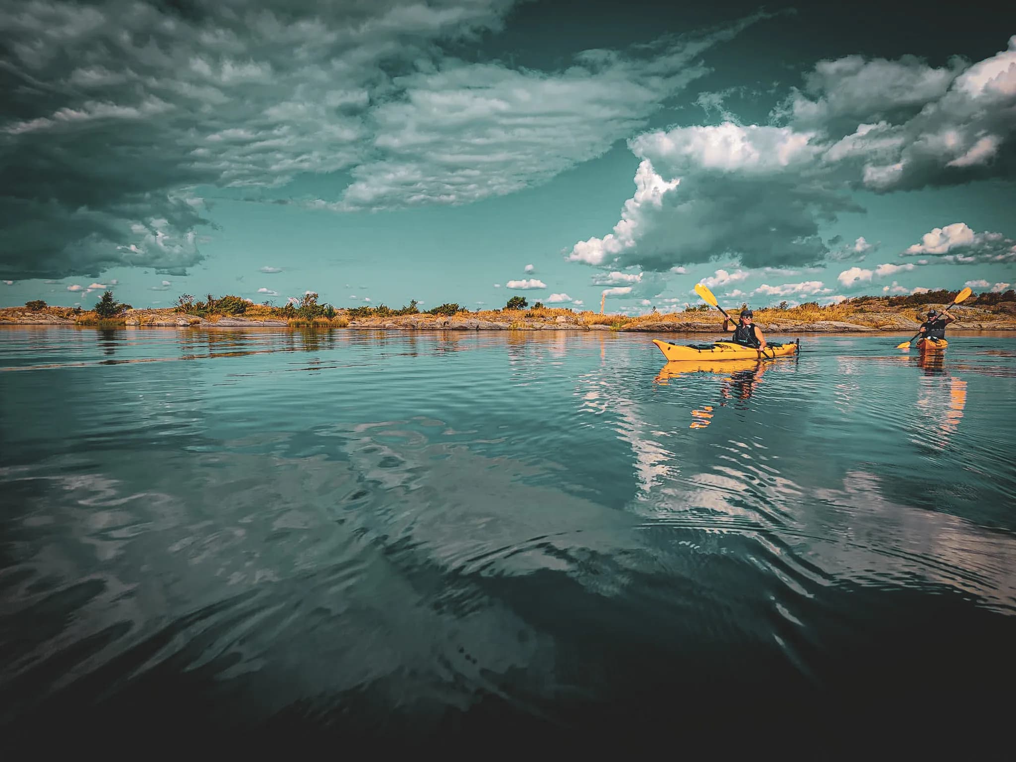 A peaceful waterscape with calm waters reflecting the cloudy sky. Two people in yellow kayaks paddled slowly, surrounded by small islands and low vegetation. The sky is bright blue with