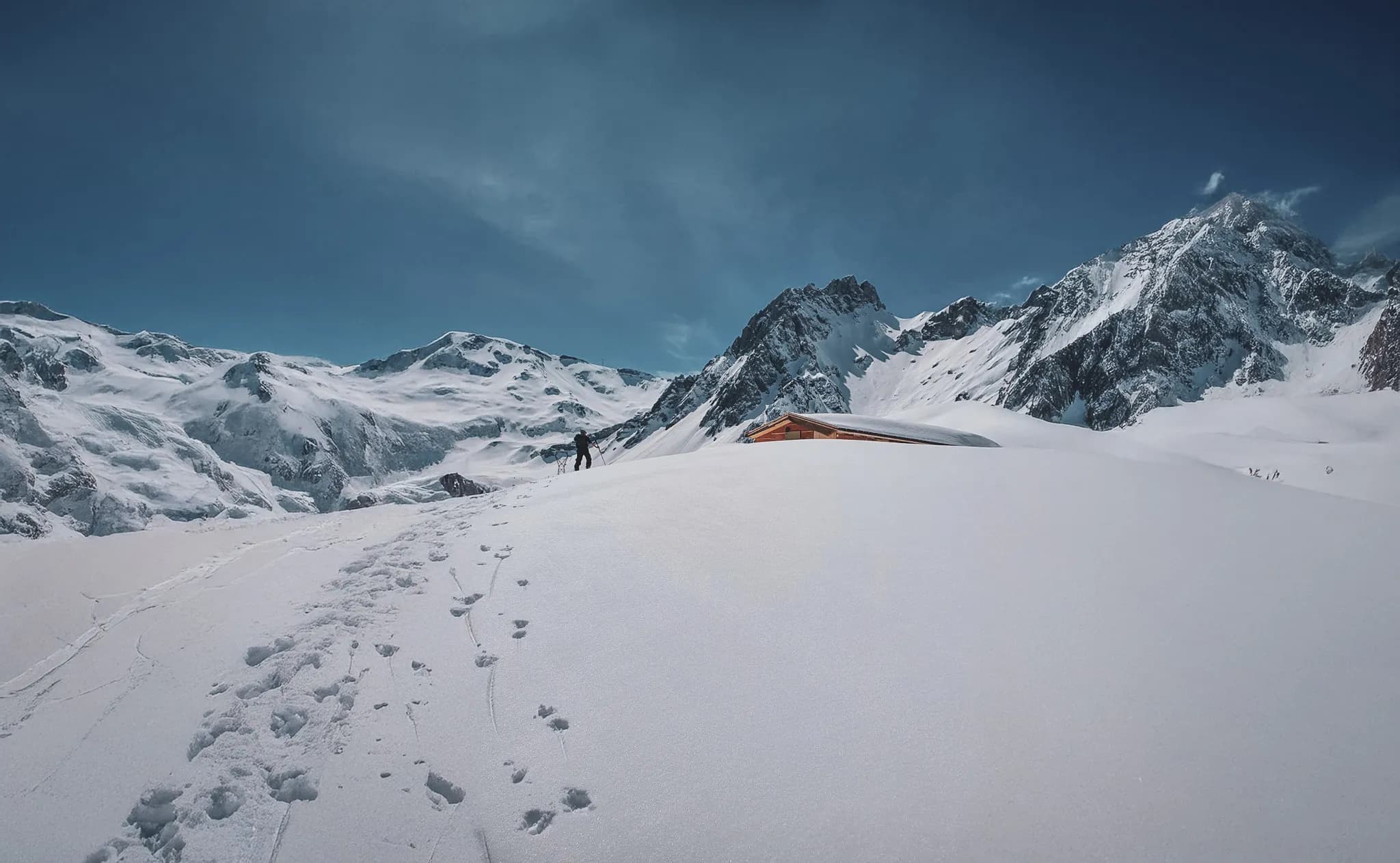Un paysage montagneux enneigé avec des sommets majestueux au loin. Au premier plan, une étendue de neige immaculée est marquée par des traces de pas. Vers la droite, un petit chalet en bois se dresse,