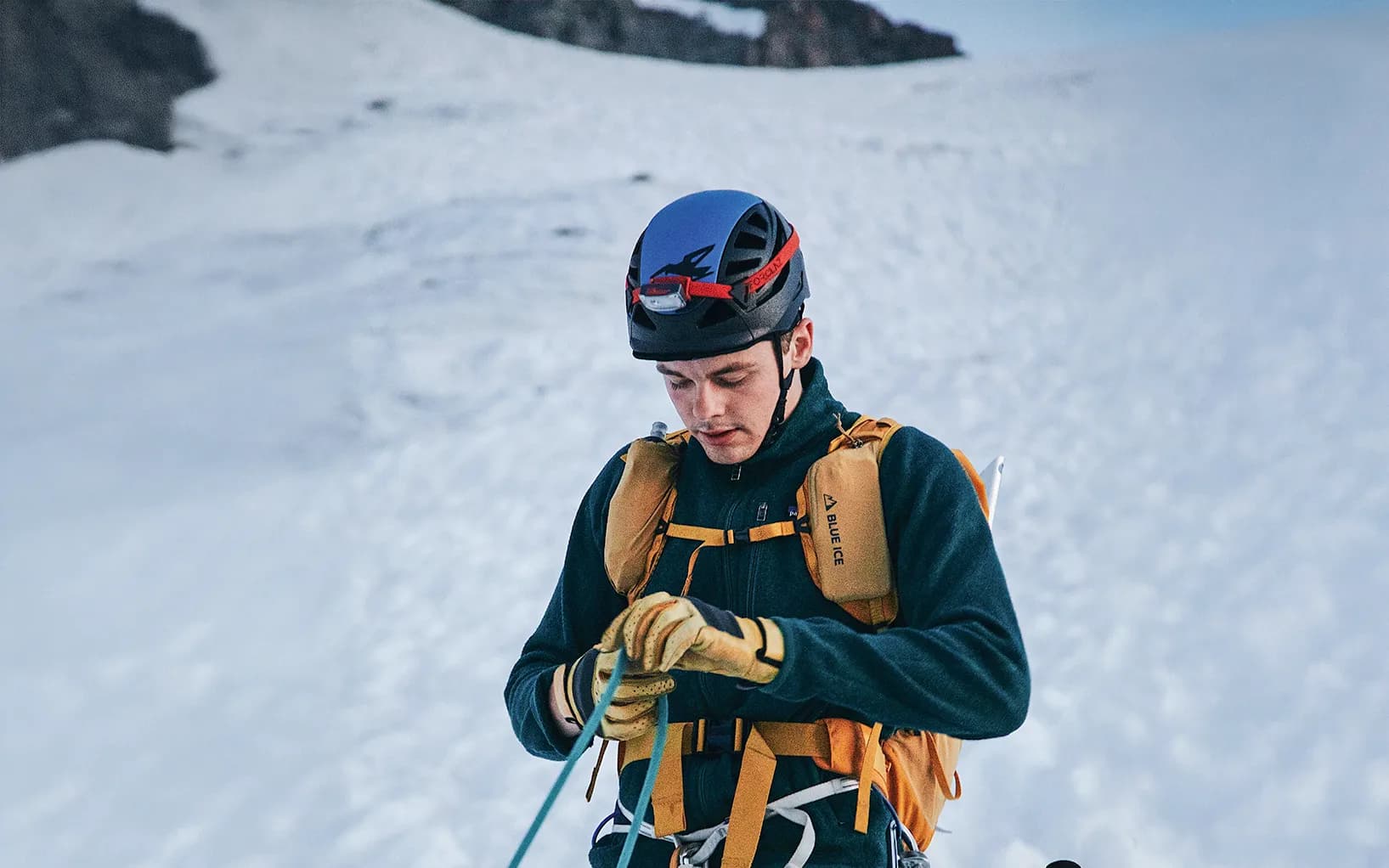 A mountaineer in full concentration, wearing a helmet and safety harness, adjusts a rope while standing on a glacier. He is wearing yellow gloves and a dark jacket, while a vast landscape of snow stretches out before him.