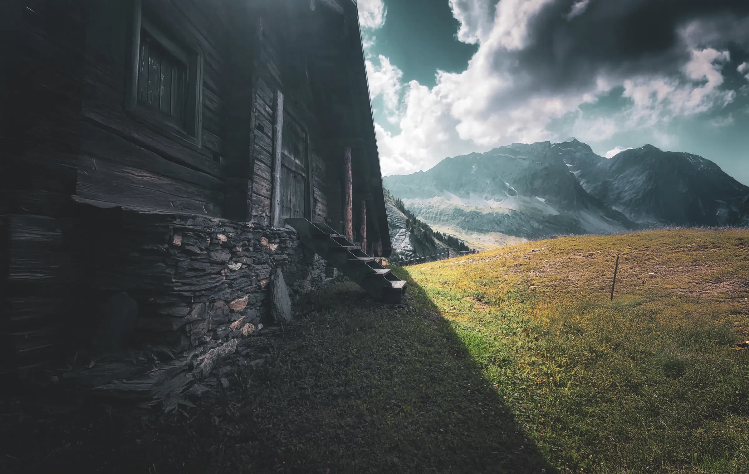 A wooden chalet sits on the edge of a green meadow, with steps leading up to a wooden door. The building is partially shaded, and the rustic architecture is enhanced by the textures of the walls.