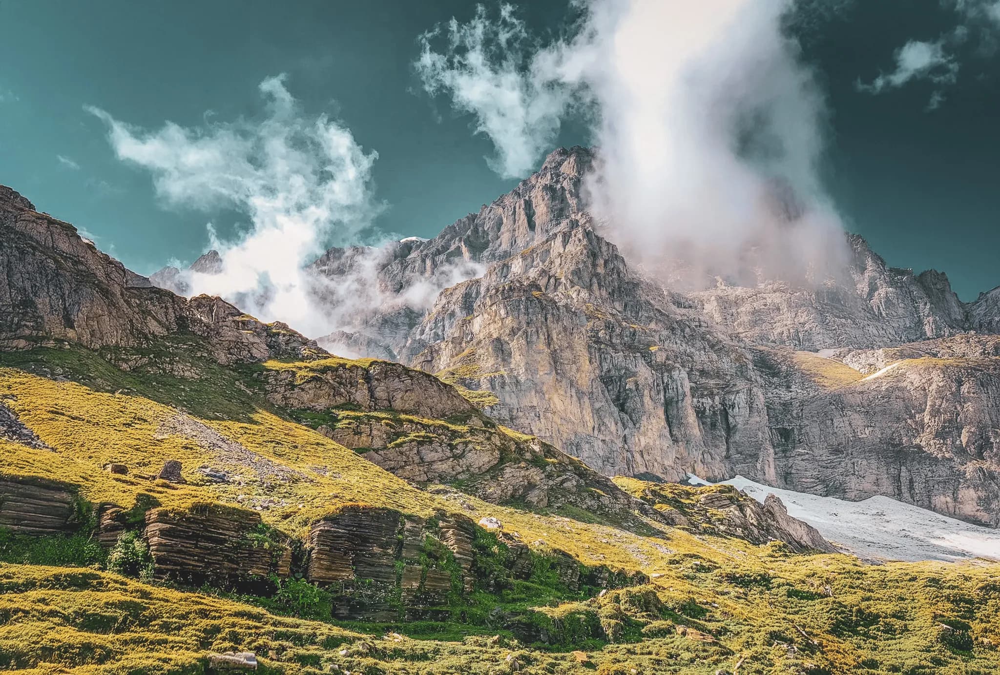A mountainous landscape with towering cliffs, whose peaks are shrouded in cloud. The base of the mountains is covered in lush greenery and grassy meadows, contrasting with the